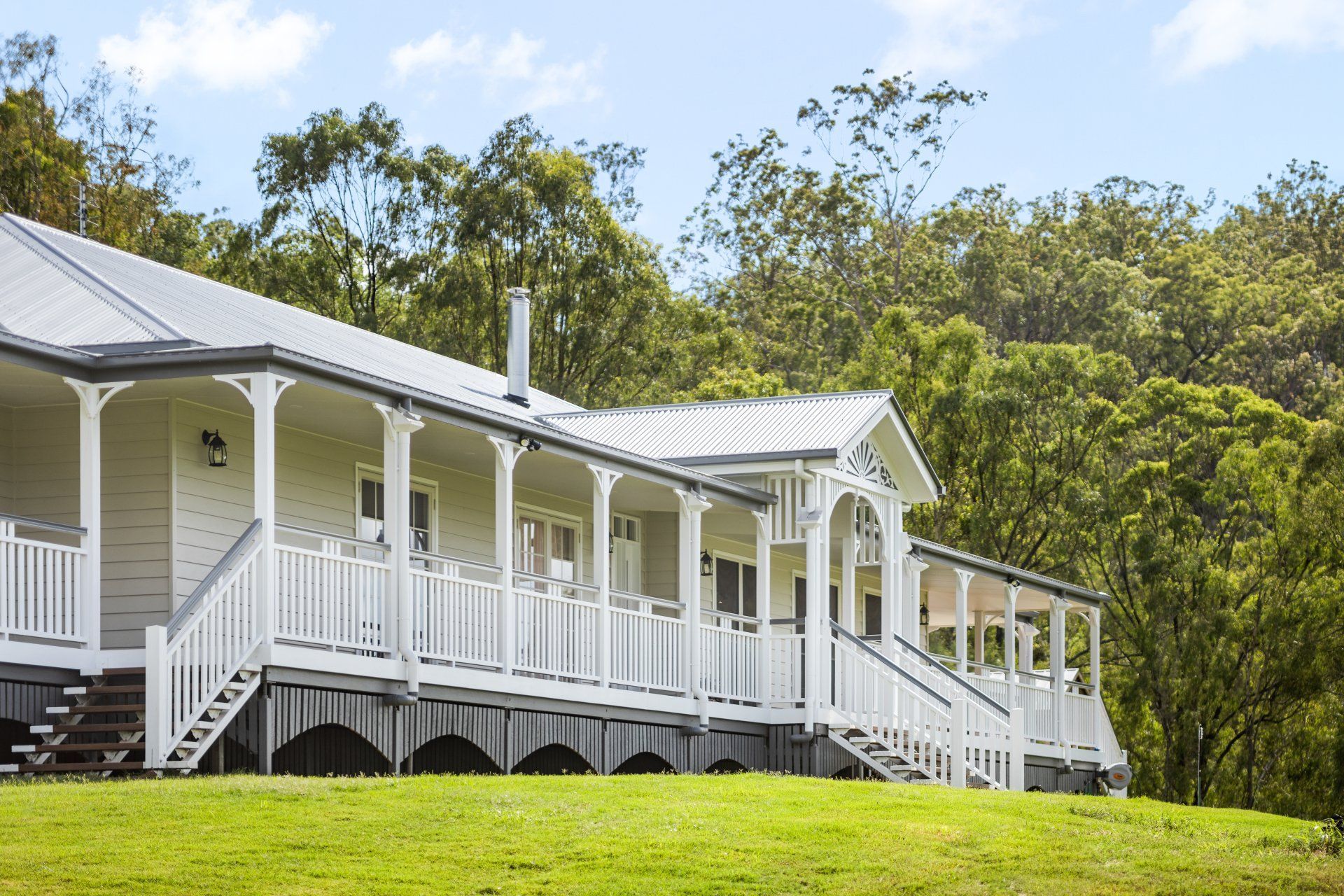 White house with wrap-around porch, on a grassy hill, backed by trees, under a blue sky.