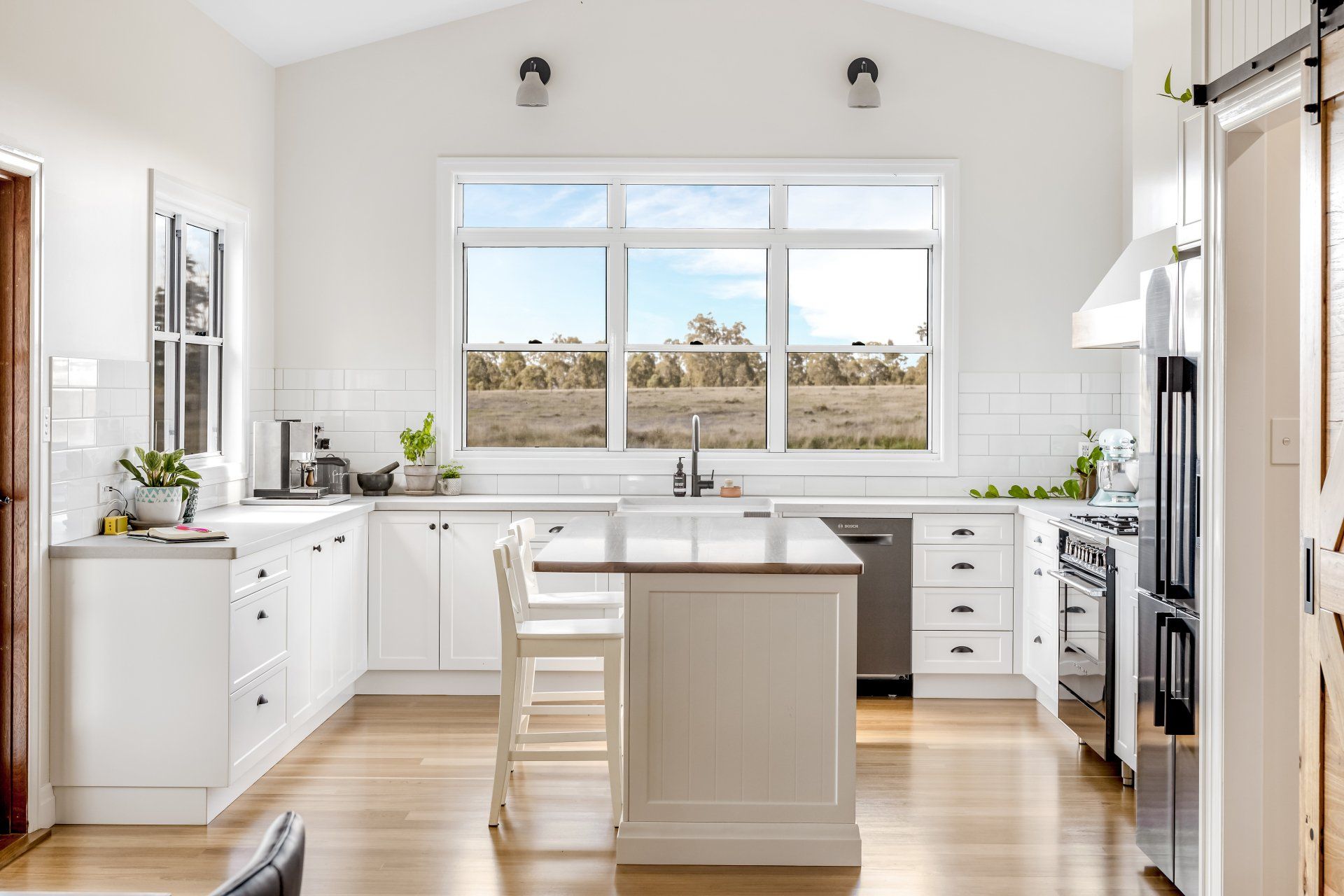 White kitchen with island, large window overlooking a field, stainless steel appliances, and wood floors.