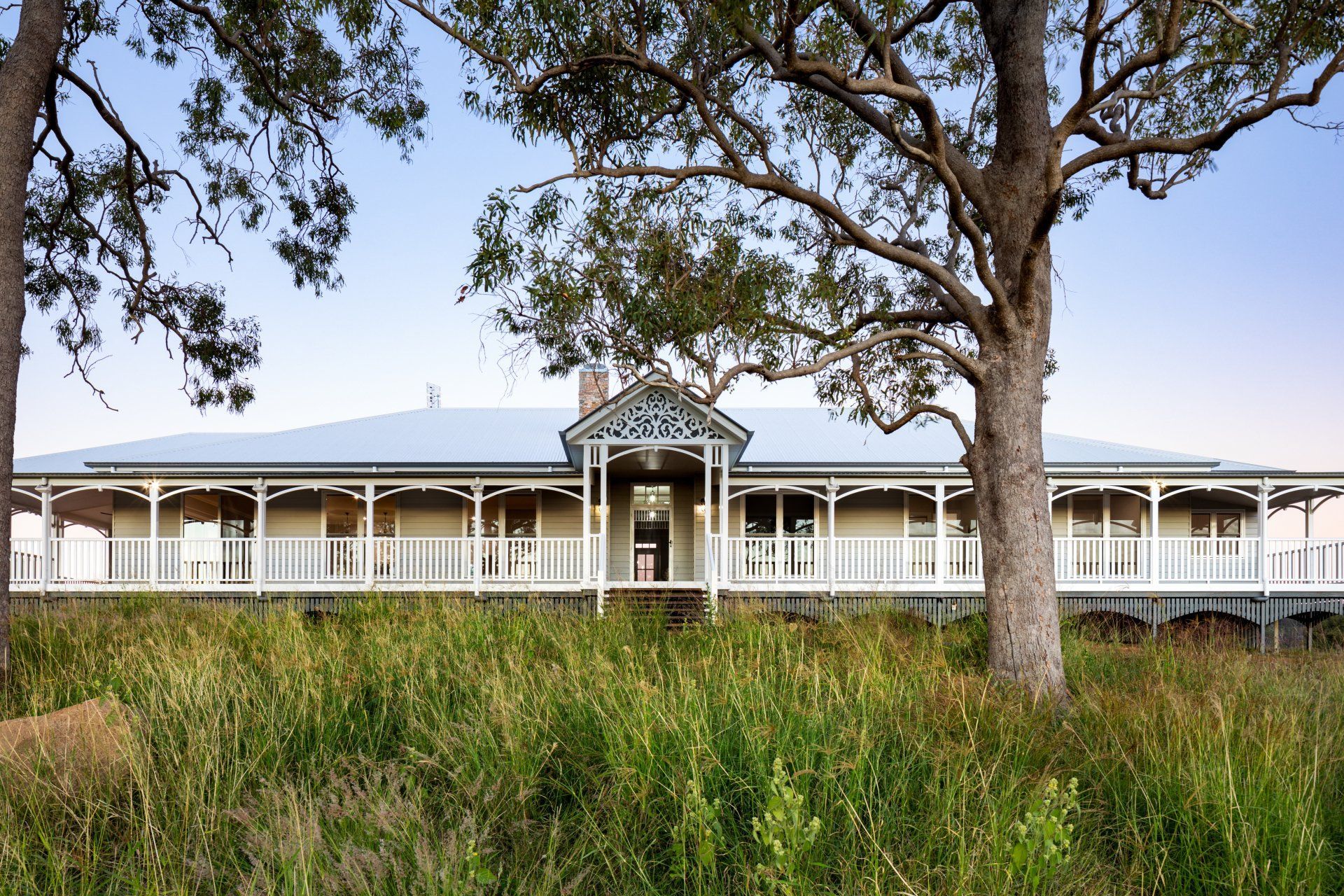 Long, light-colored house with wrap-around porch, framed by trees and tall grass.