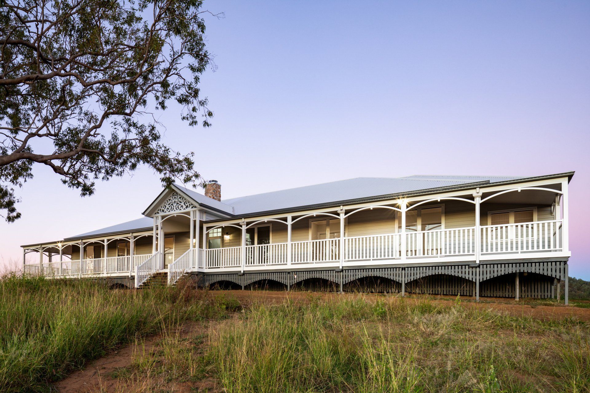 A large, light-colored house with a wraparound porch in a grassy field at dusk.
