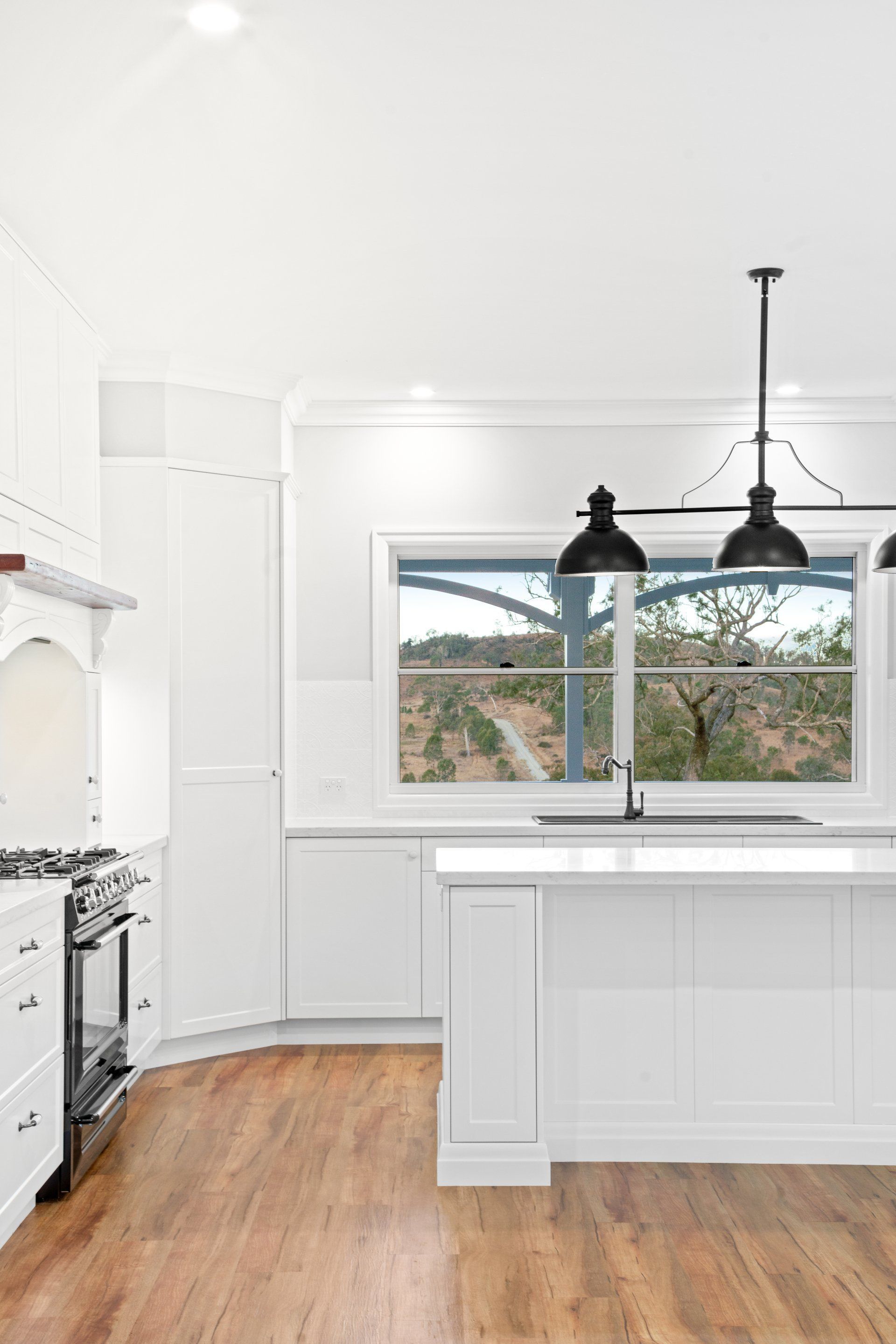 White kitchen with hardwood floors, island, black pendant lights, and a window overlooking a landscape.