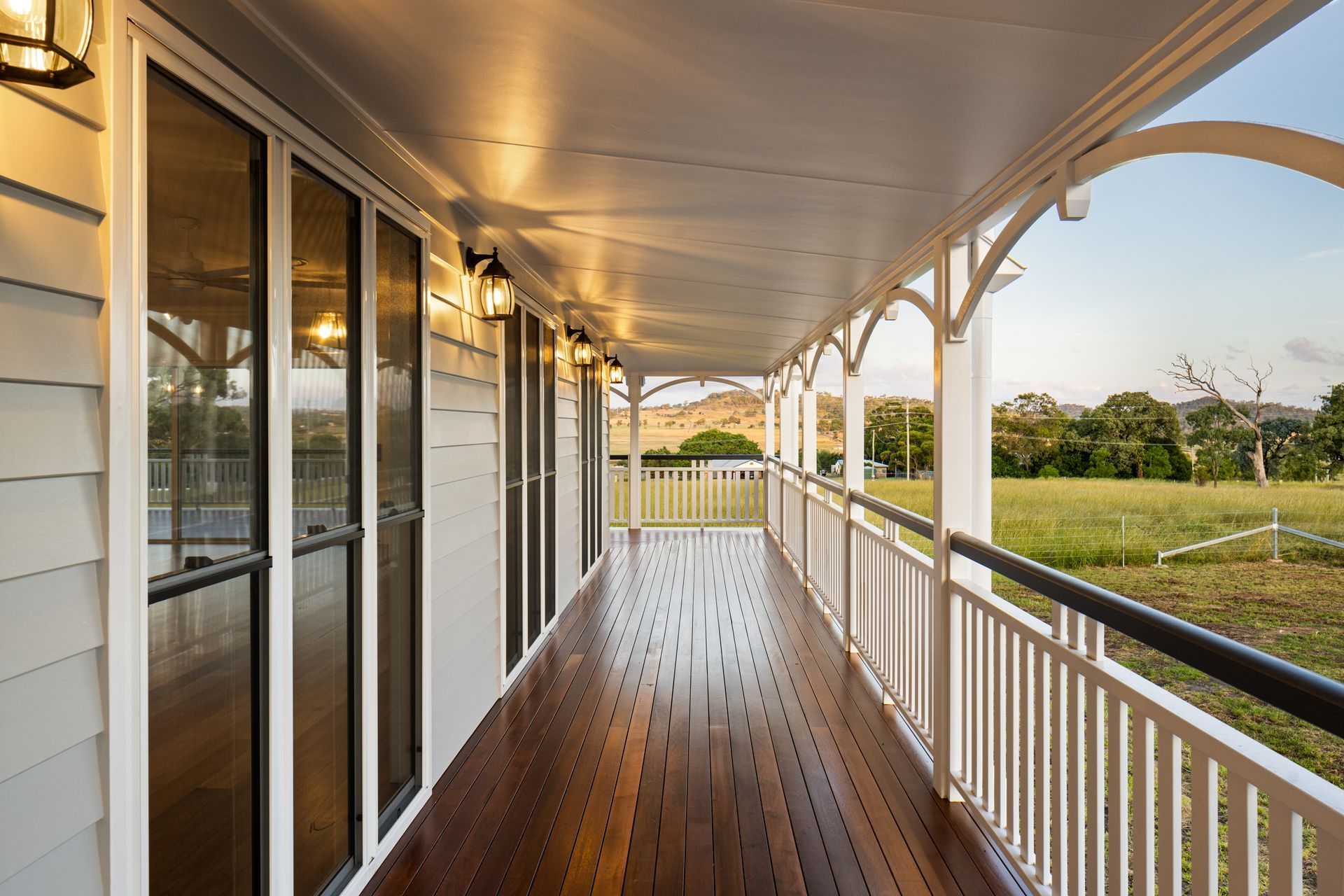 Wooden veranda with white railing and light fixtures, overlooking a grassy field.