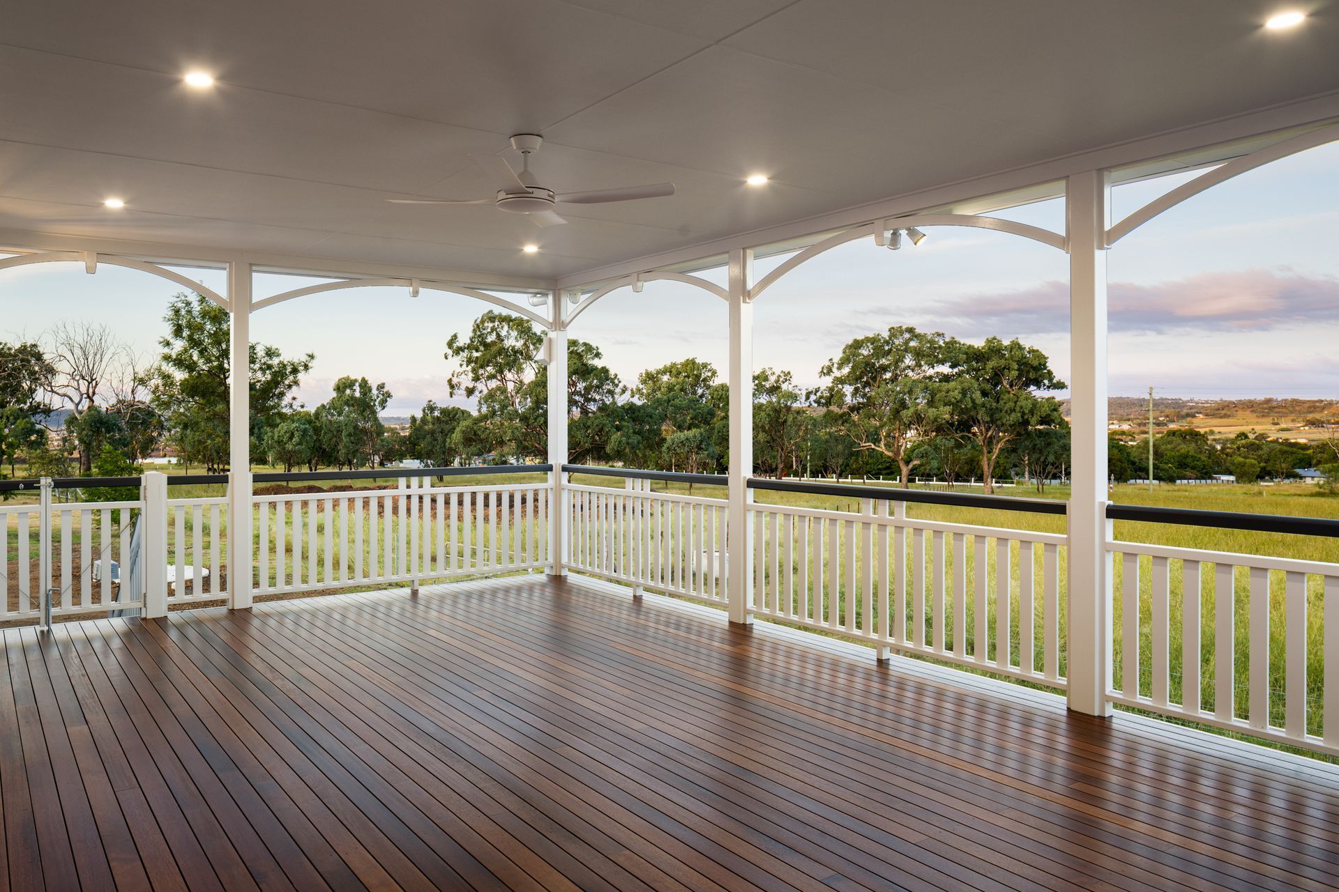 Wooden veranda with white railing and ornate archways, overlooking a green landscape.