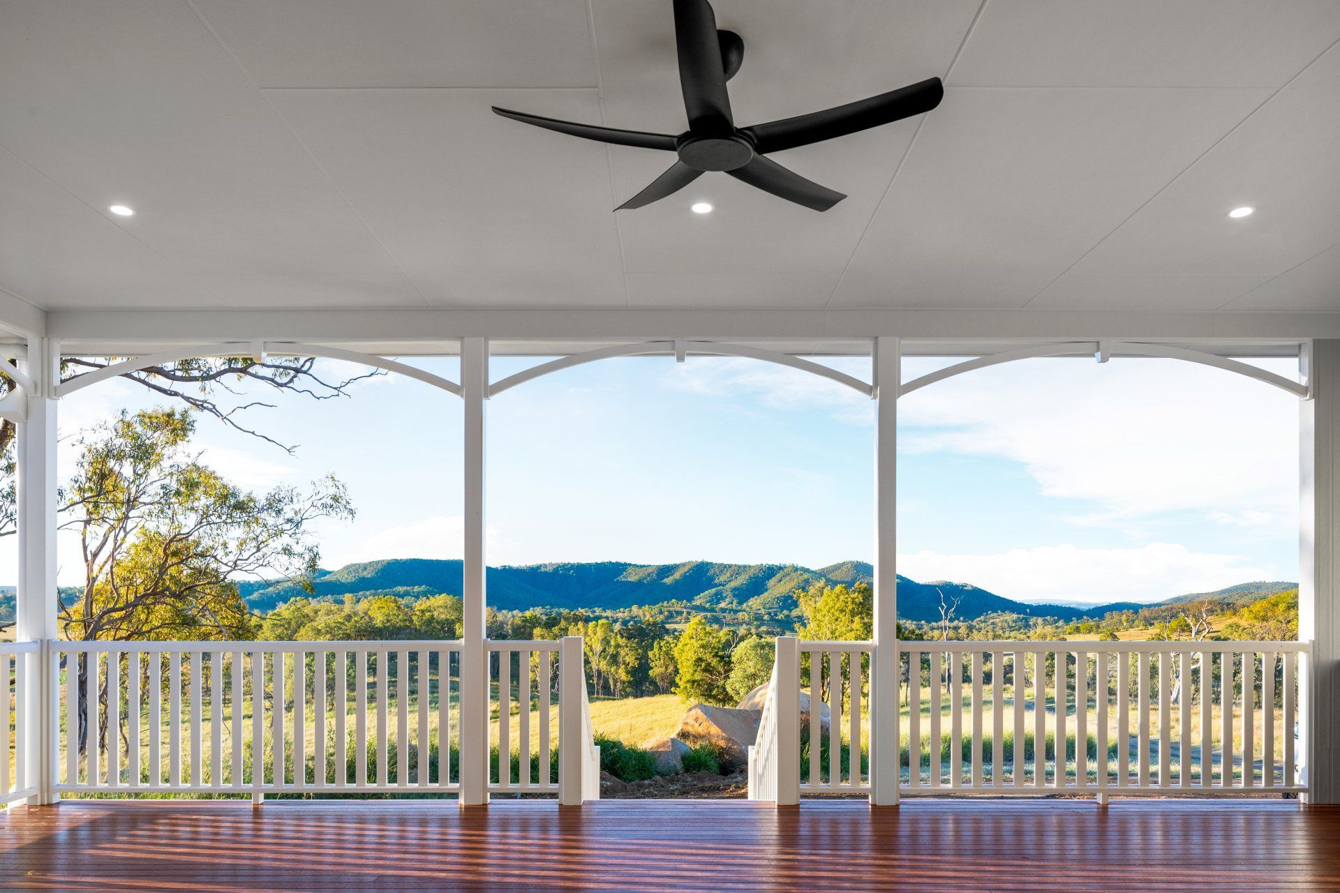 Veranda overlooking green valley, white railing, arched window frames. Ceiling fan, blue sky.