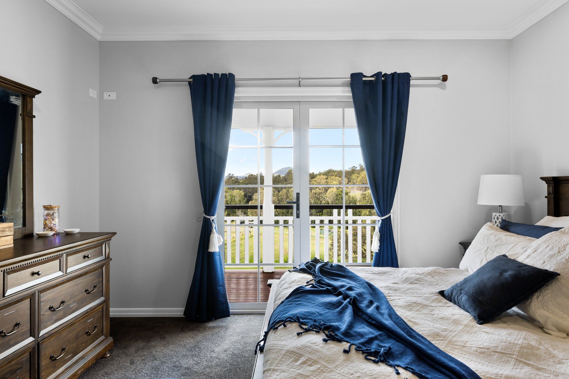 Bedroom with bed, dresser, and blue curtains framing a view of a porch and trees.