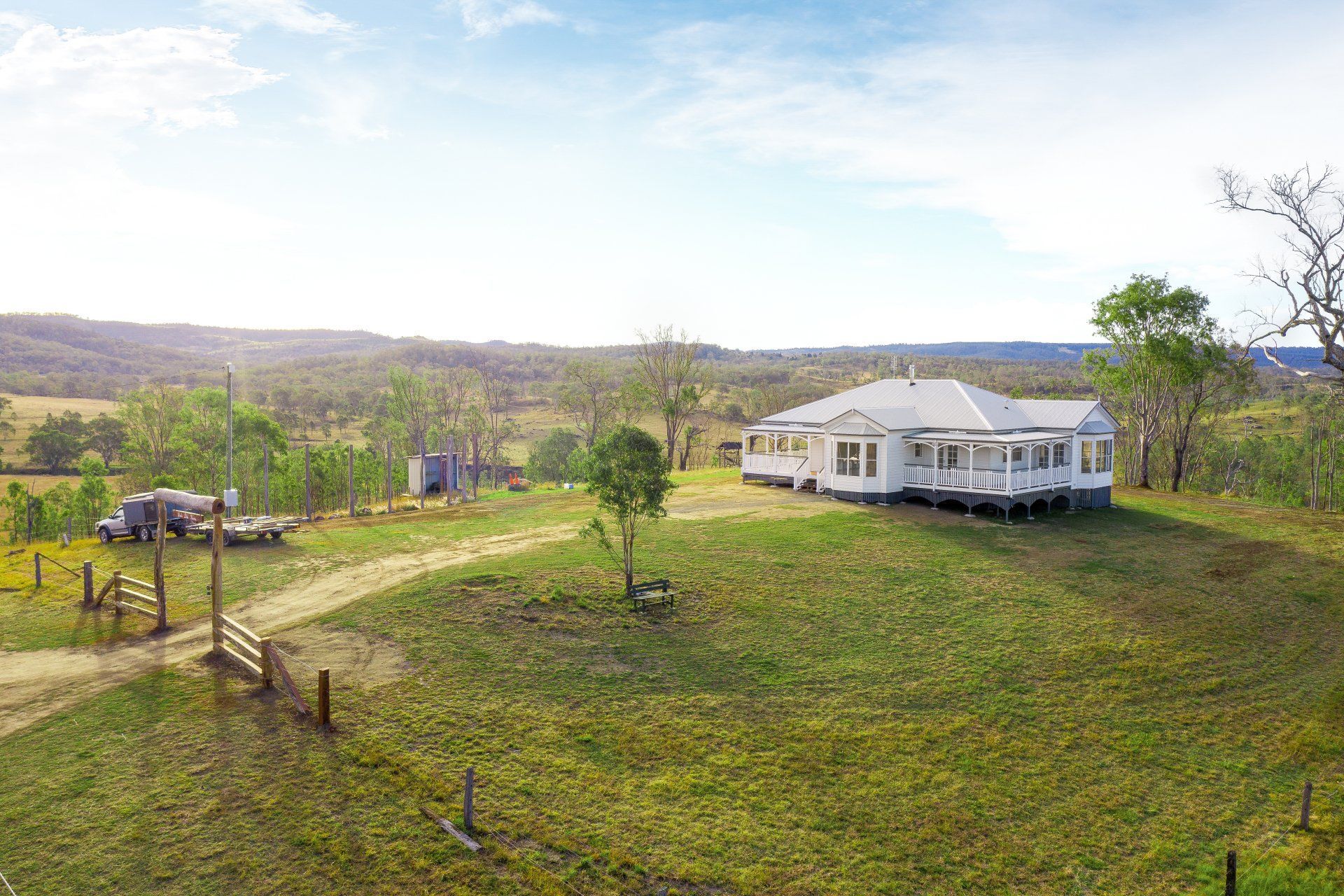 White farmhouse on a grassy hill; dirt driveway and surrounding trees. Sunny day.