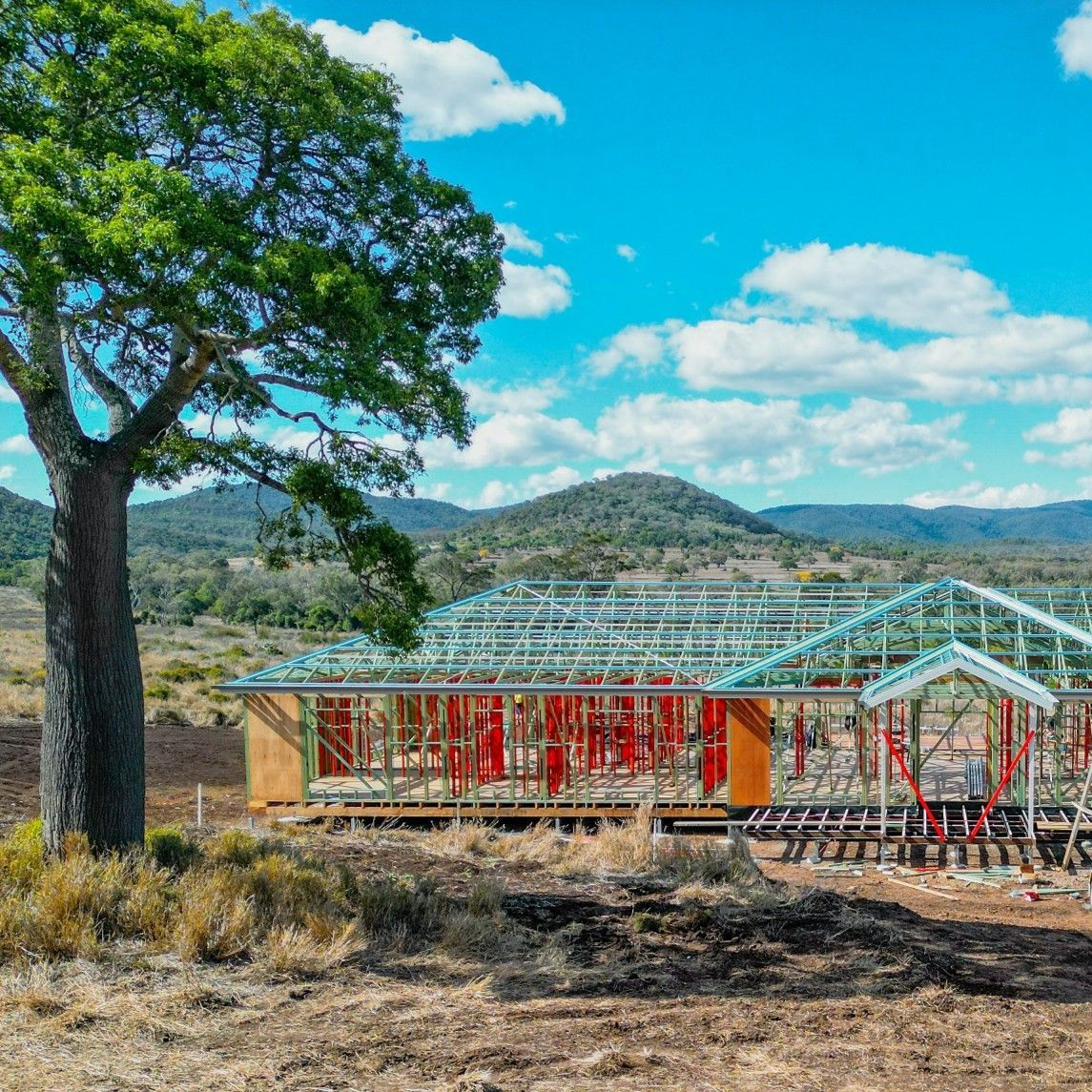 Construction of a house with metal frame under blue sky, tree in the foreground, mountains in the background.
