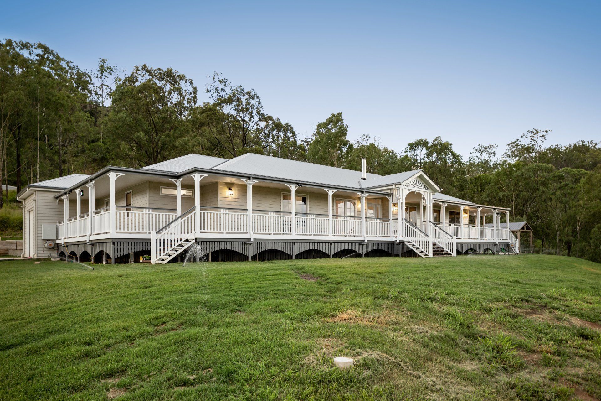 White house with wraparound porch on a grassy hill, surrounded by trees under a blue sky.