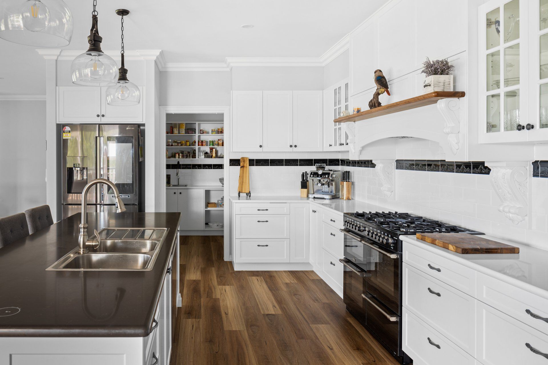 White kitchen with dark countertops and wood flooring. Stainless steel sink, black range, and pantry.