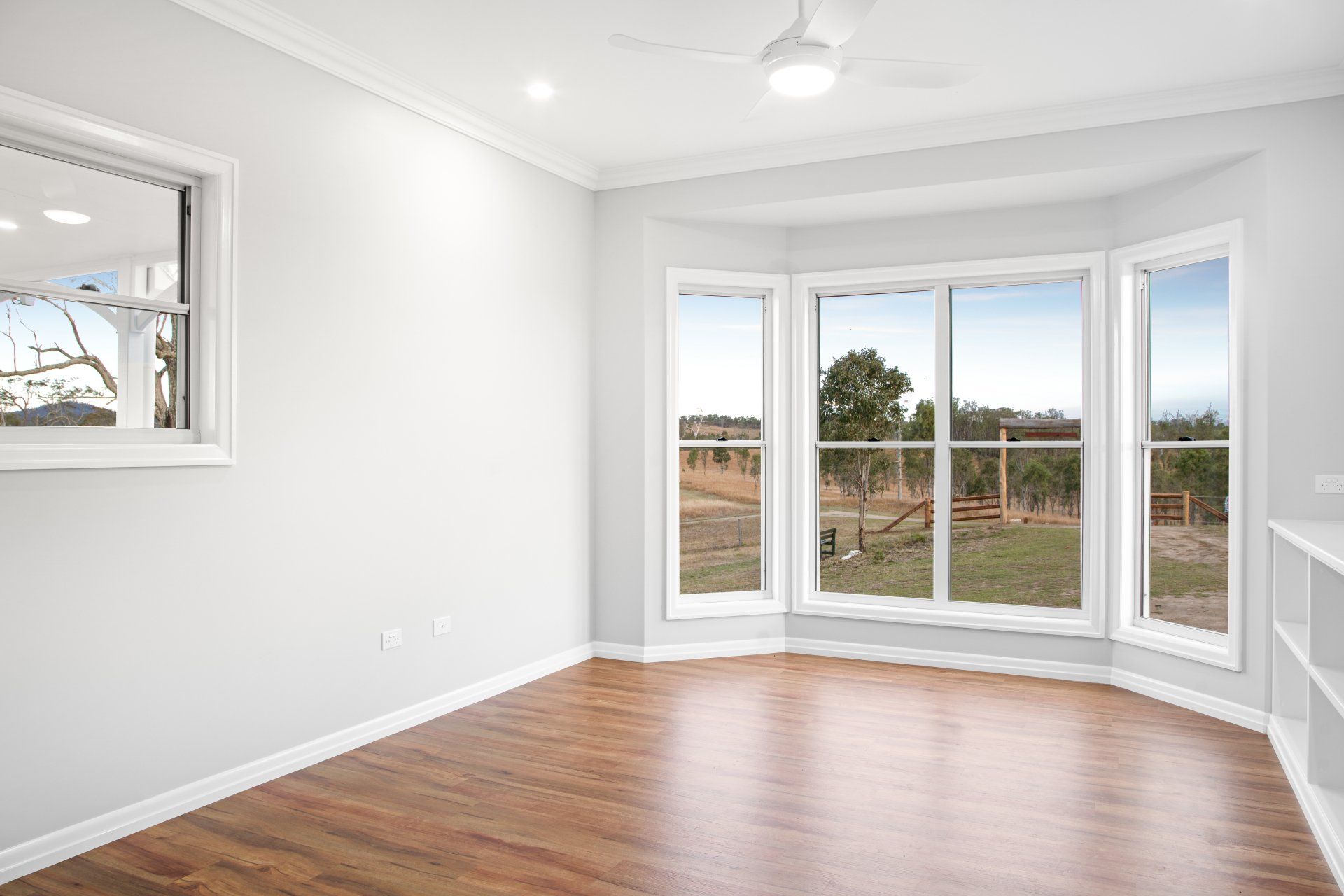 Empty room with wooden floor and large windows overlooking a field, a smaller window on the left.