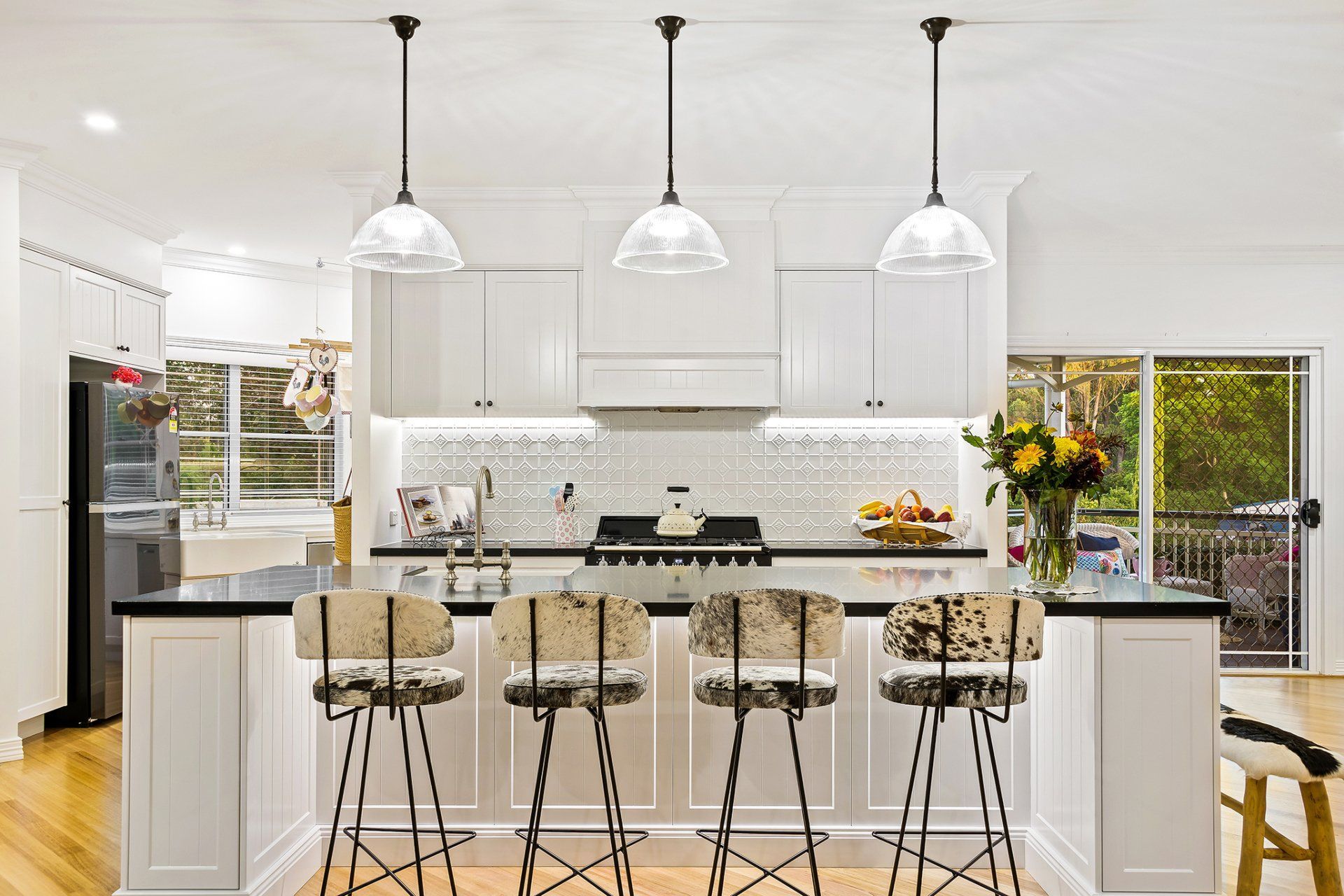 White kitchen with a black countertop island and four cowhide bar stools. Three pendant lights hang above.