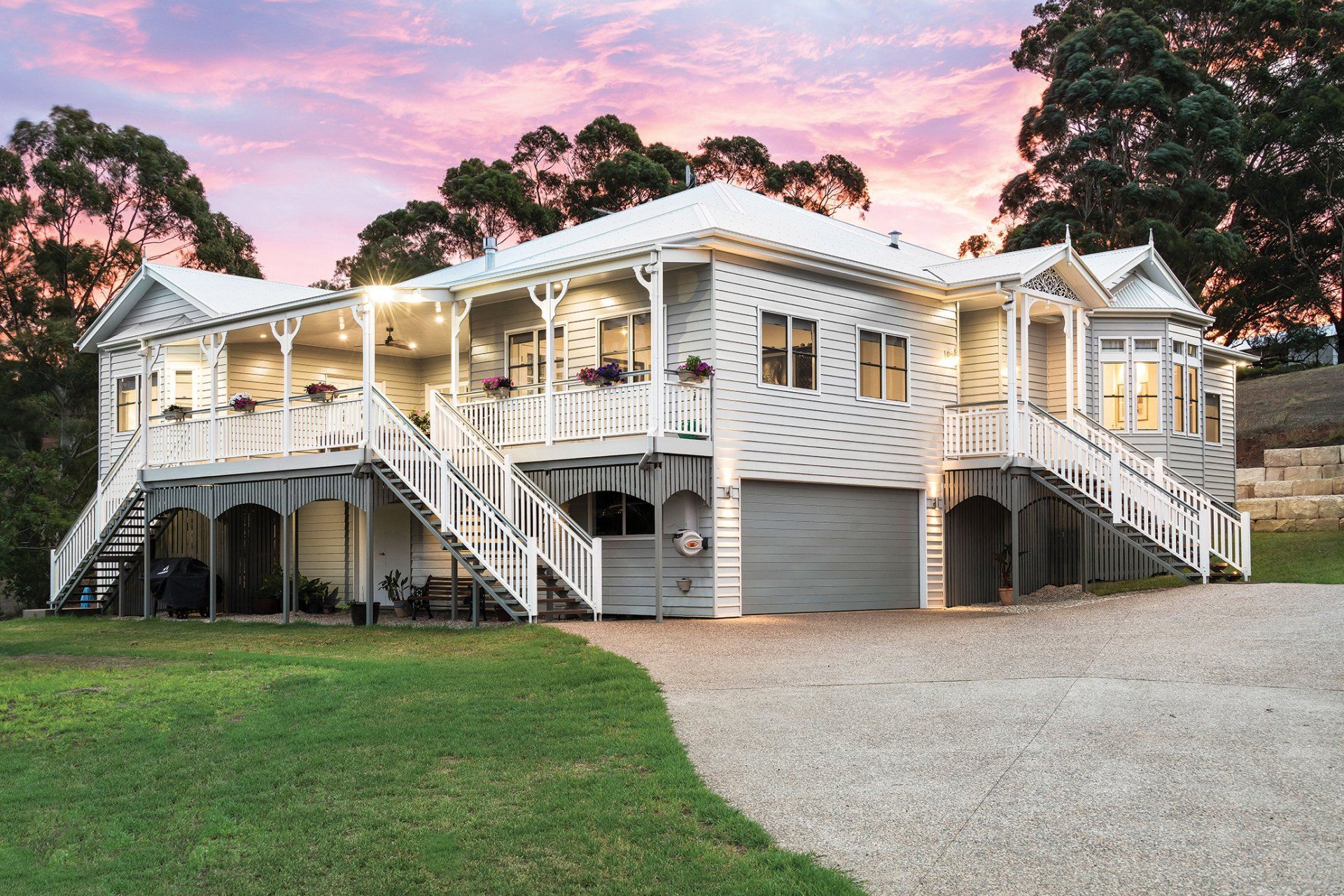 White two-story house with wrap-around porch, gray garage door, and gravel driveway. Pink and purple sunset.