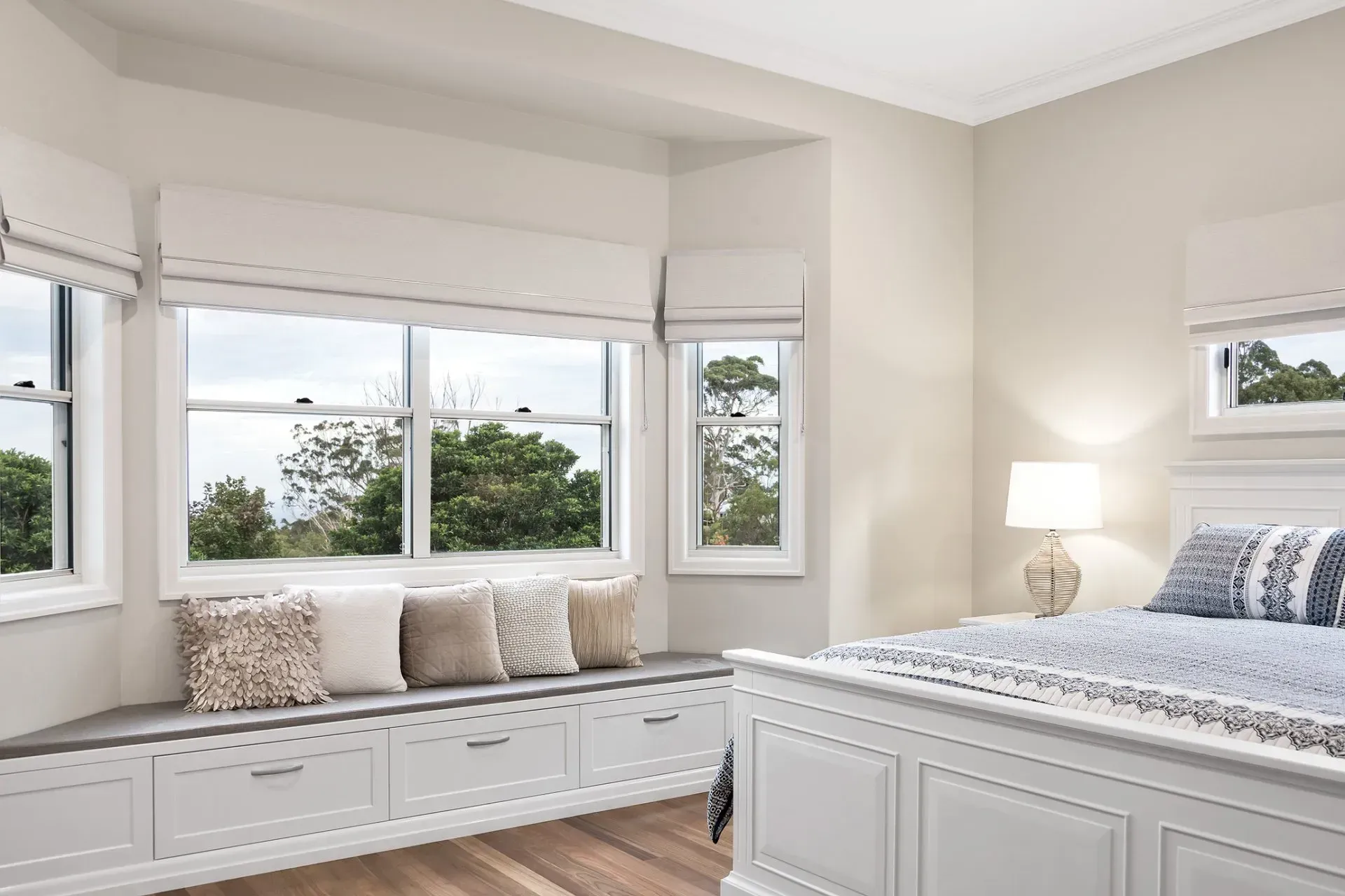 Bedroom with window seat, white bed, neutral walls, and roman shades.
