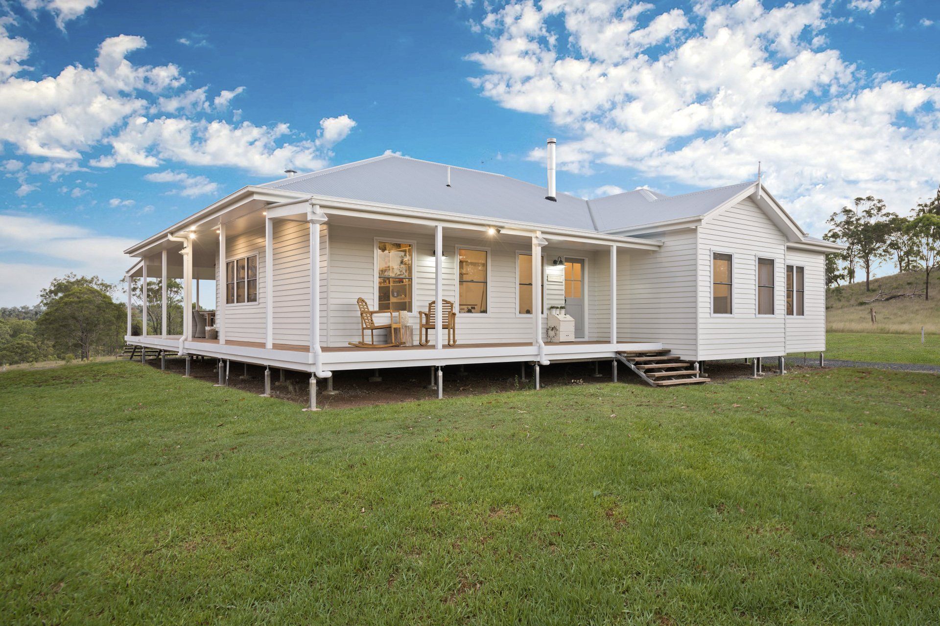 White farmhouse with a wraparound porch, set on a grassy hill under a blue sky with clouds.