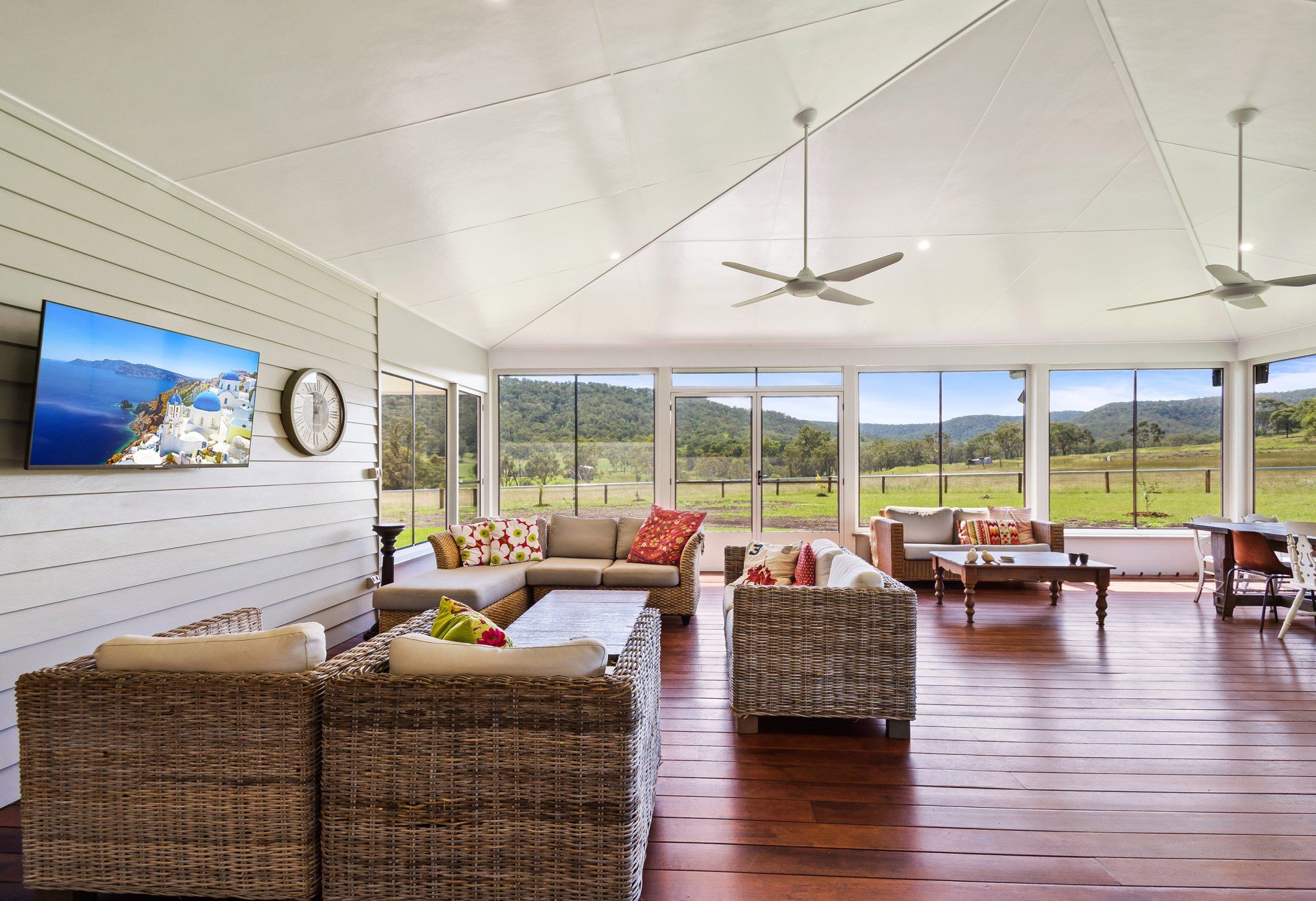 Spacious living room with large windows overlooking a green field. Multiple couches, TV, ceiling fans, and wood floor.