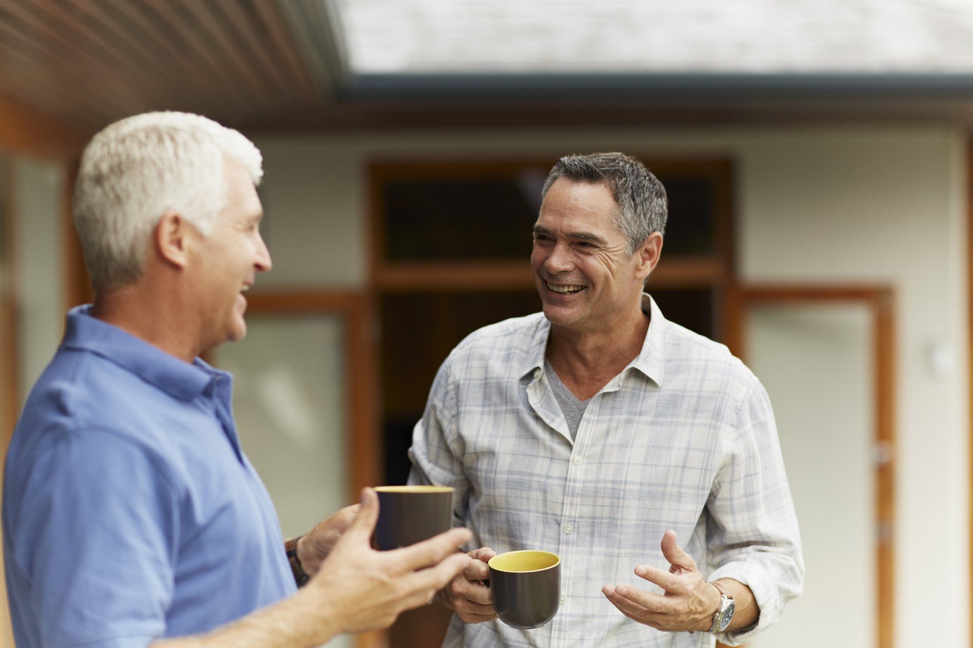 Two men, outdoors, conversing and smiling while holding mugs.