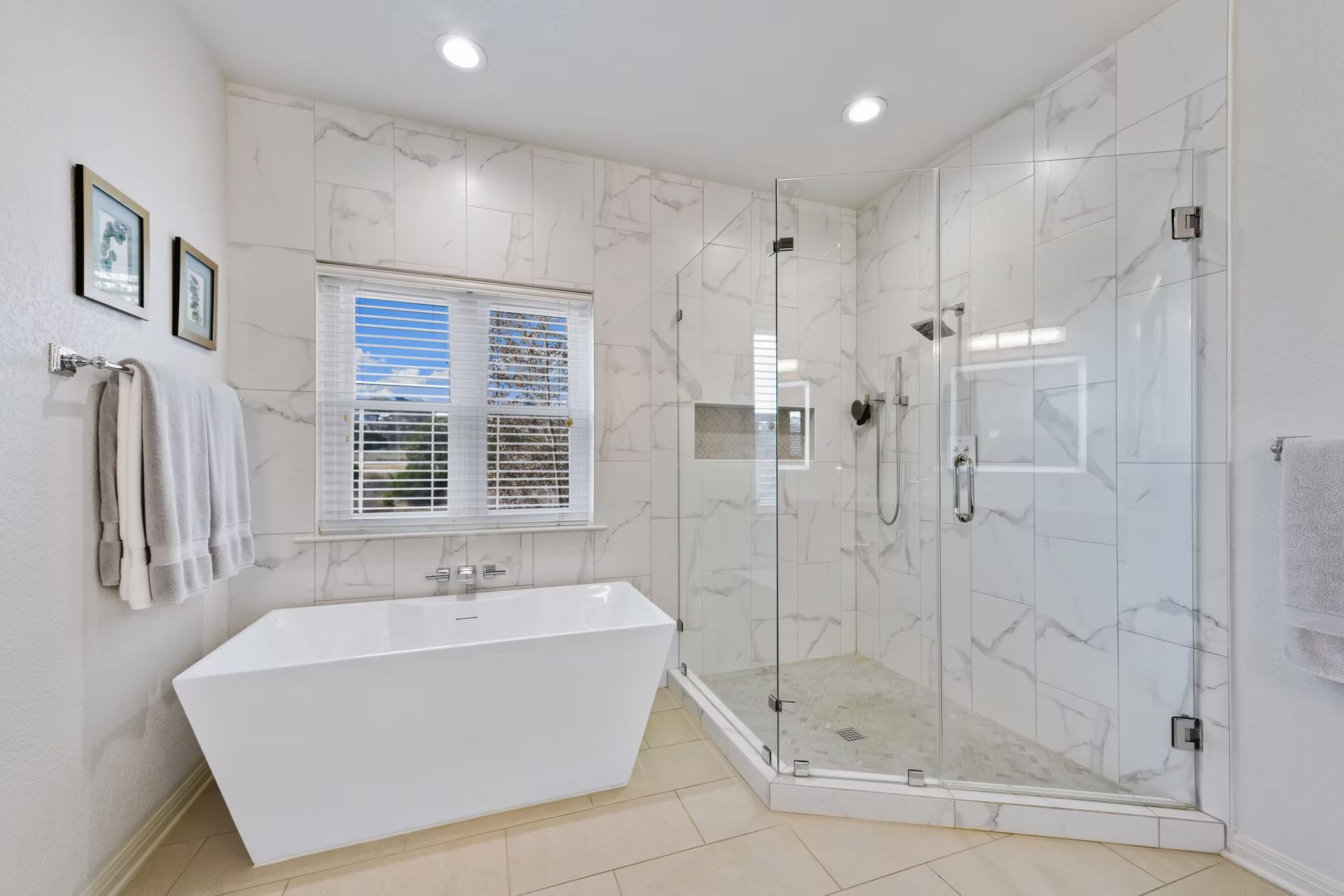 Bright marble bathroom with a freestanding tub, glass shower, window, and white towels