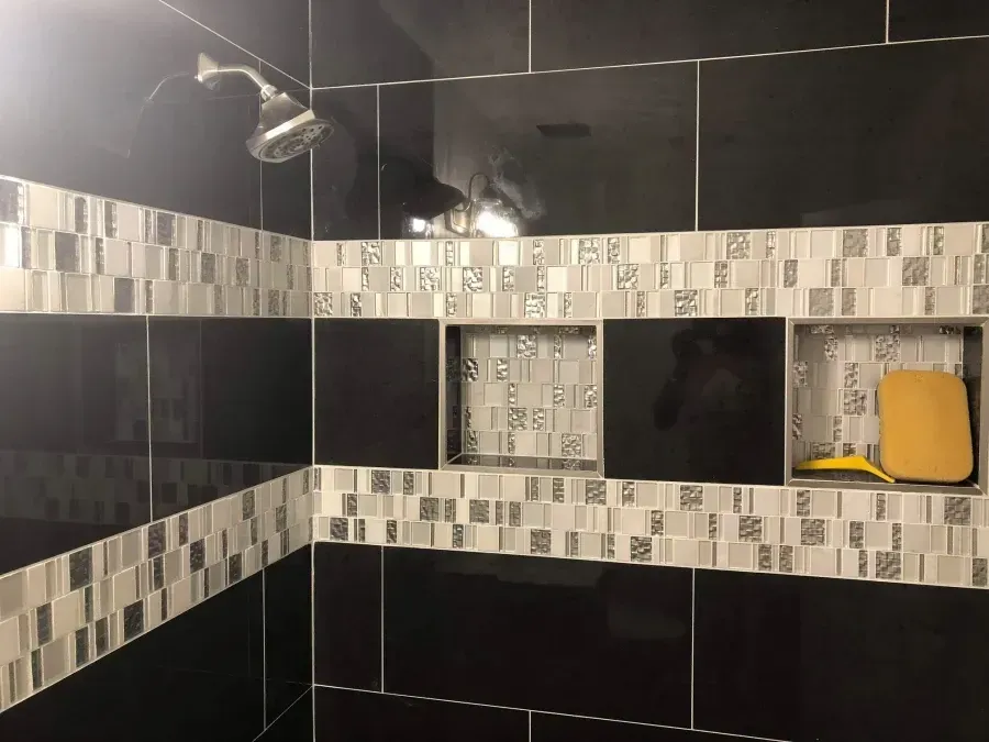 Modern bathroom shower with black tile and gray mosaic trim, featuring a showerhead and built-in shelf with a yellow item