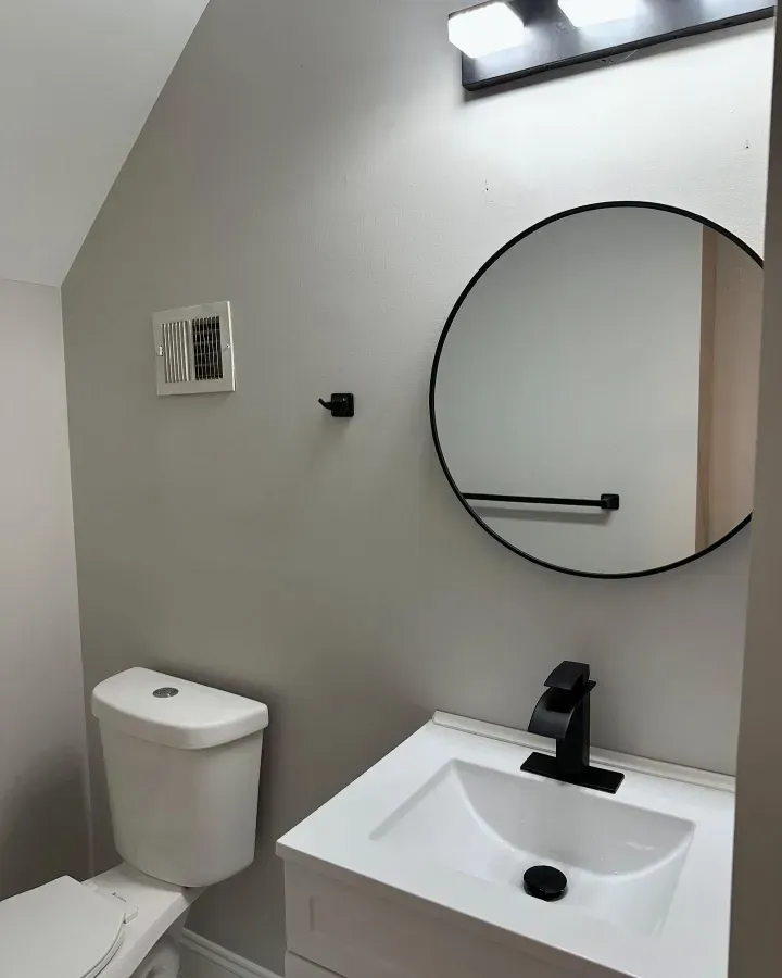 Small bathroom with toilet, white sink, round mirror, and wall light on a gray wall.