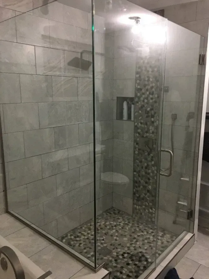 Glass walk-in shower with gray tile walls and pebble-stone floor, viewed from the bathroom.