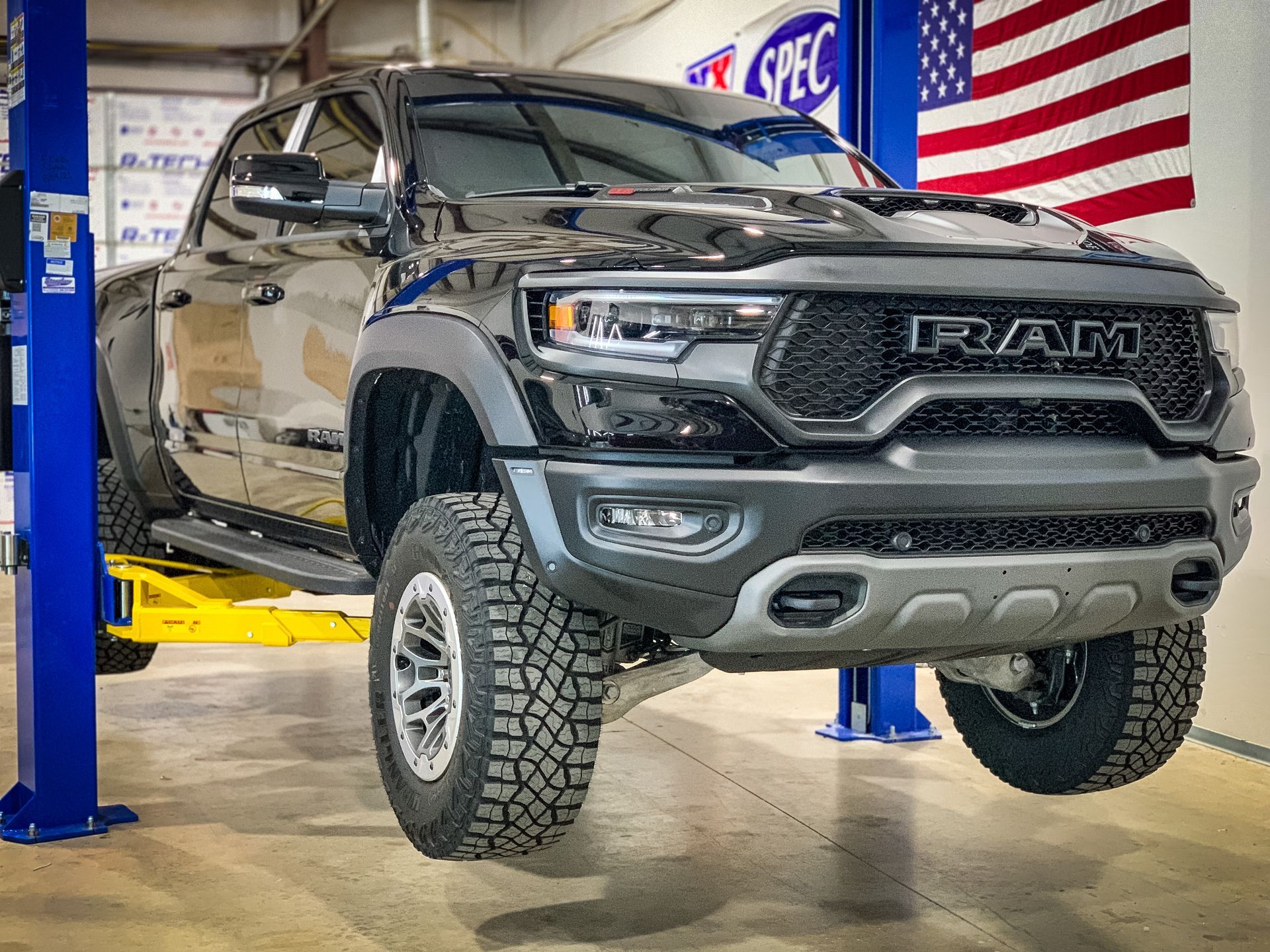 Black RAM TRX truck on a car lift in a shop, American flag in background.