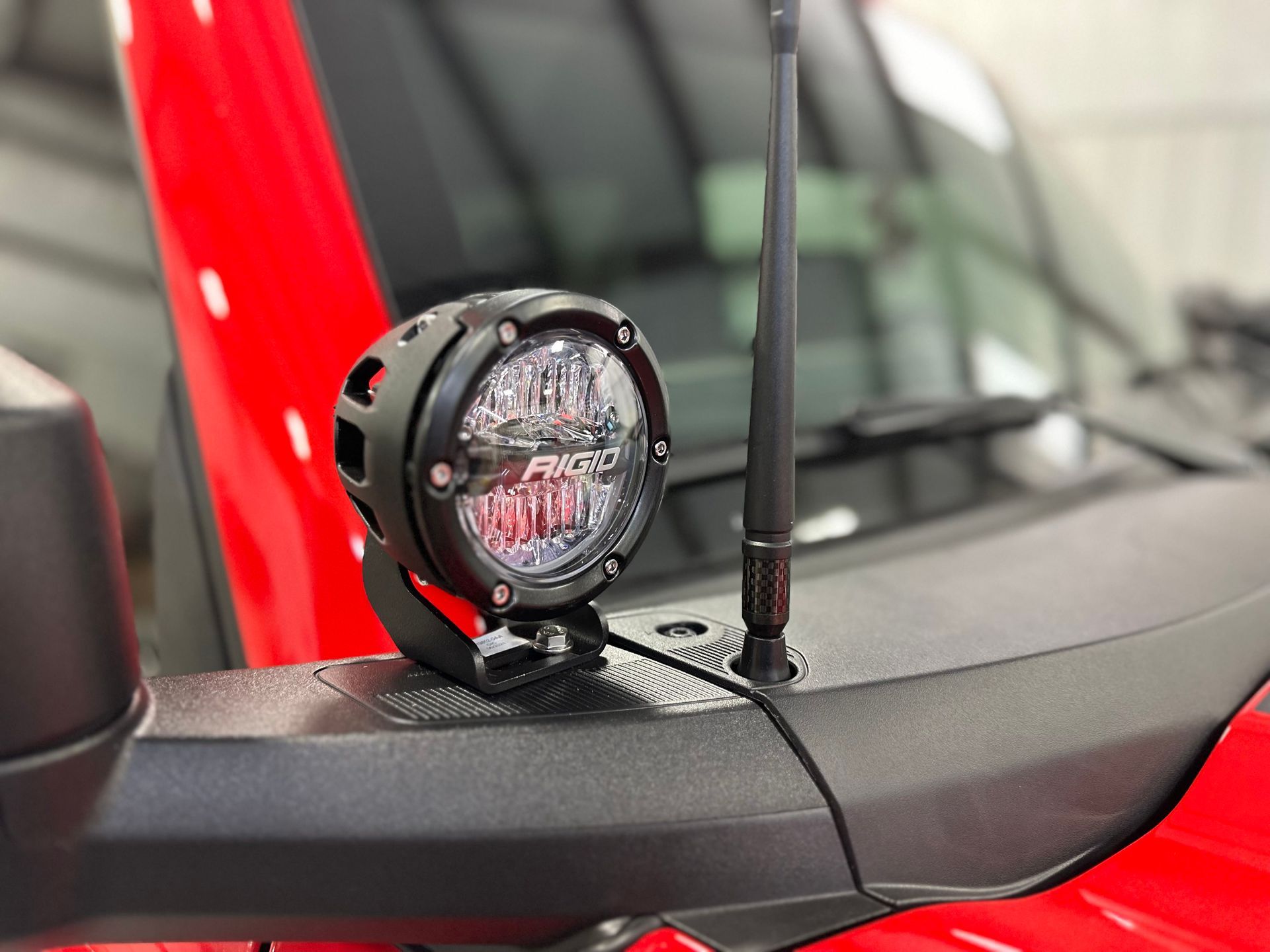Black LED spotlight mounted on a red vehicle's hood next to a black antenna.