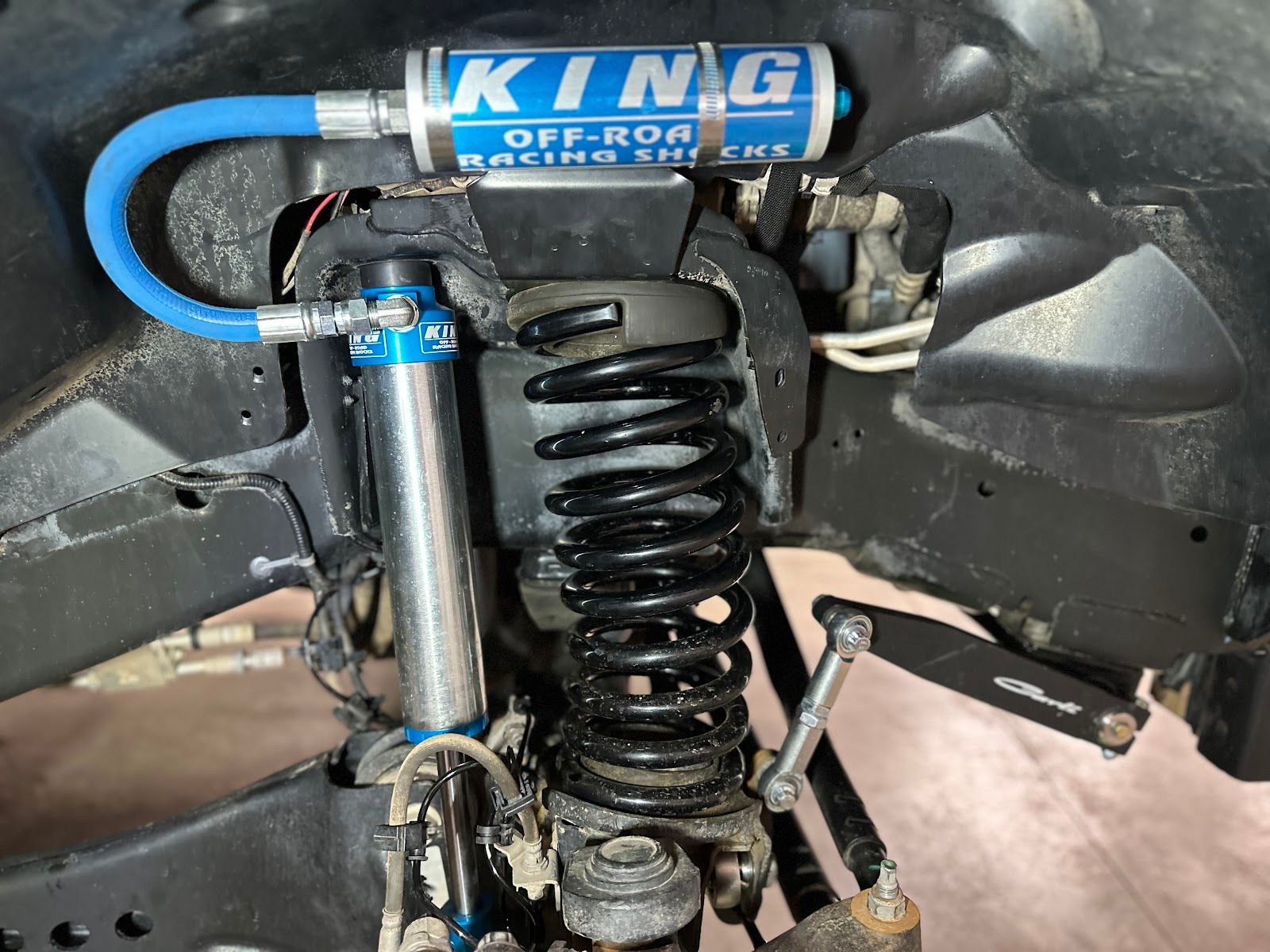 A close-up of a Jeep suspension featuring King off-road shocks and a blue reservoir, connected by a blue hose.