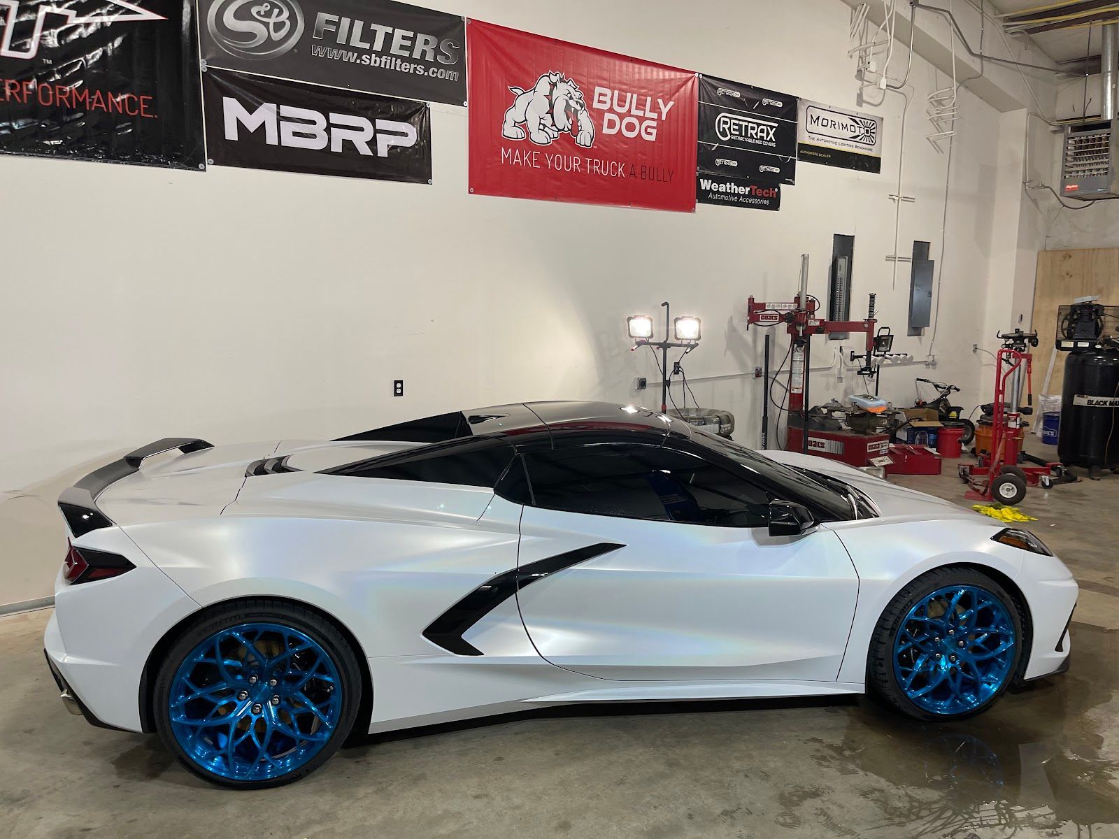 White Corvette with blue rims parked inside a garage. Banners for car parts are visible in the background.