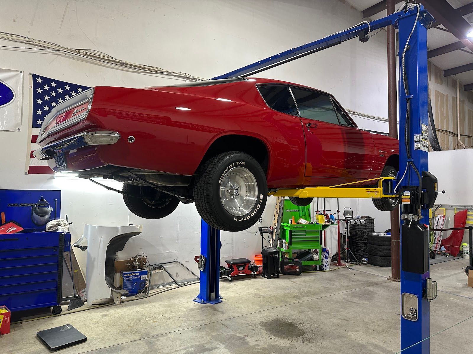 Red classic car raised on a blue and yellow lift in a garage, with tools and an American flag visible.