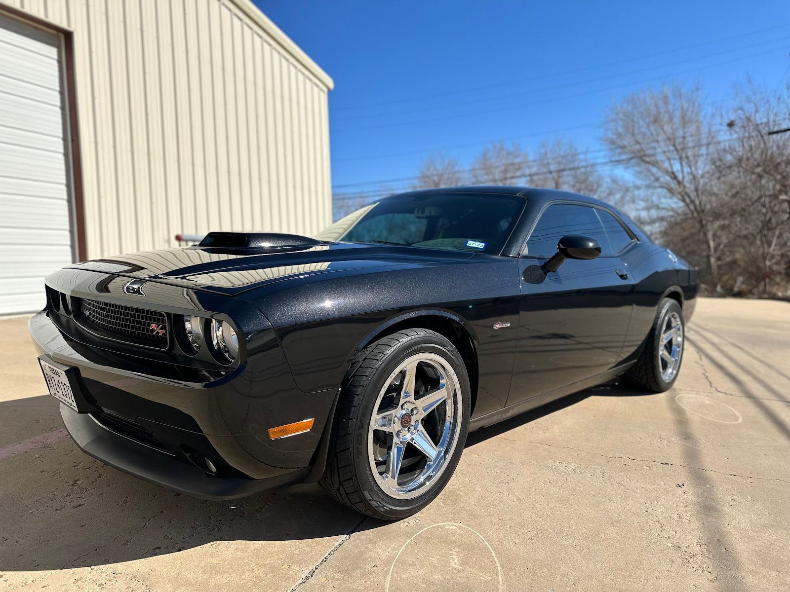 Black Dodge Challenger sports car parked in front of a building on a sunny day. It has chrome wheels and a hood scoop.