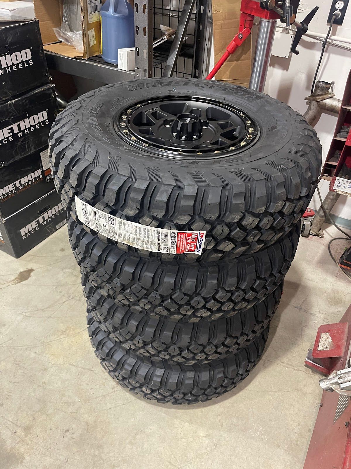 Stack of four black off-road tires with black rims. They are in a garage or shop setting, with boxes and tools visible.