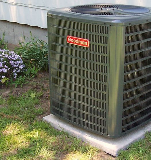 Goodman air conditioner unit on a concrete pad near a building, surrounded by grass and plants.