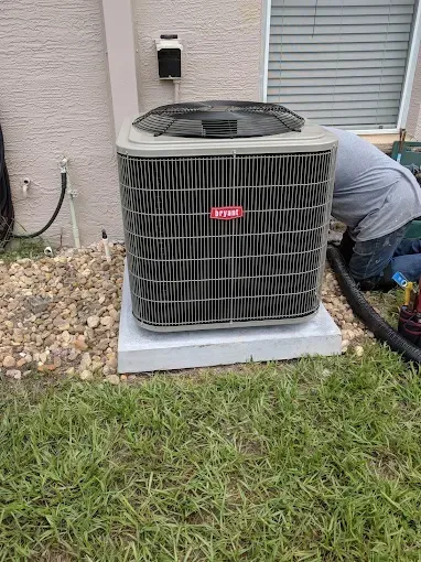 Air conditioning unit on a concrete pad, technician working on it near a building with gravel and grass.