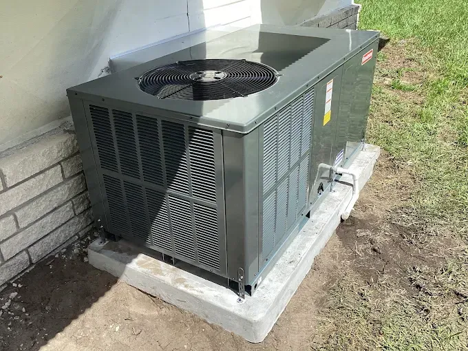 Gray air conditioning unit on a concrete pad next to a brick wall and grass.