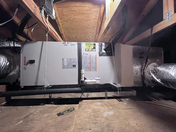 HVAC system in an attic. Rectangular grey unit with ductwork and wooden beams.