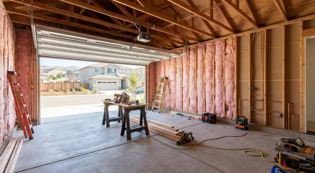 Unfinished garage interior with exposed framing, tools on the floor, and daylight opening to the outside.
