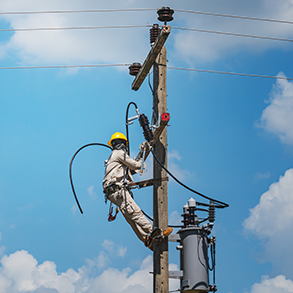 Lineman in safety gear on utility pole, working on electrical wires against a blue sky.