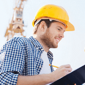Construction worker in yellow hard hat, smiling, taking notes on a clipboard in front of a crane.