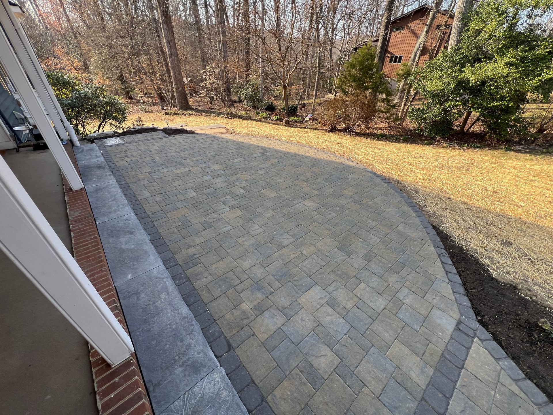 Brick patio with pavers, bordered by dark grey blocks, leading to a yard with bare trees.