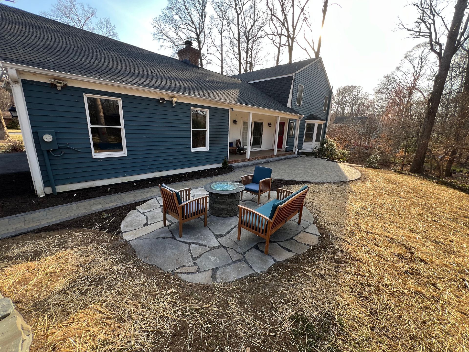 Blue house with stone patio, fire pit, and wooden chairs in a yard with brown mulch and trees.