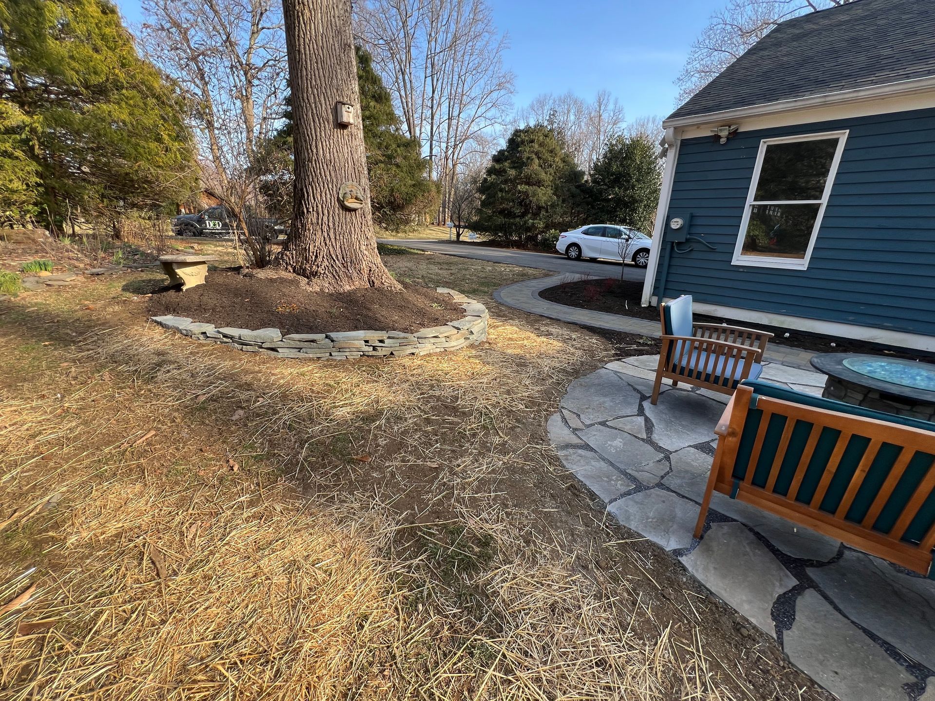 Backyard scene with a tree, a blue house, a stone patio, and two wooden benches.