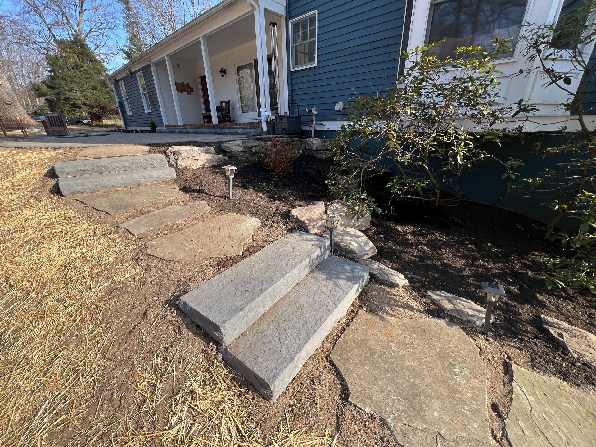 Stone steps and pathway leading to a blue house with a porch. Landscaping includes mulch and small plants.