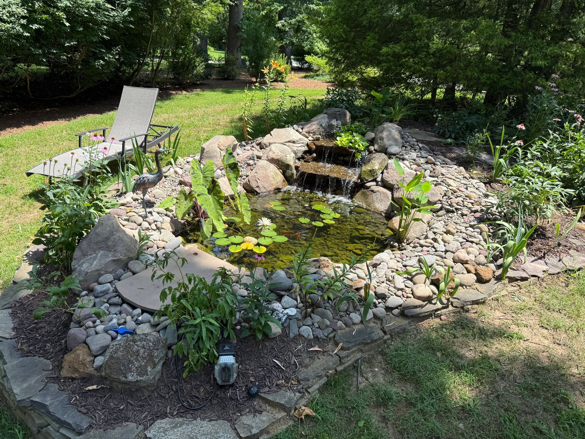 Small pond with waterfall, surrounded by rocks and plants in a yard.  An empty chair sits nearby.