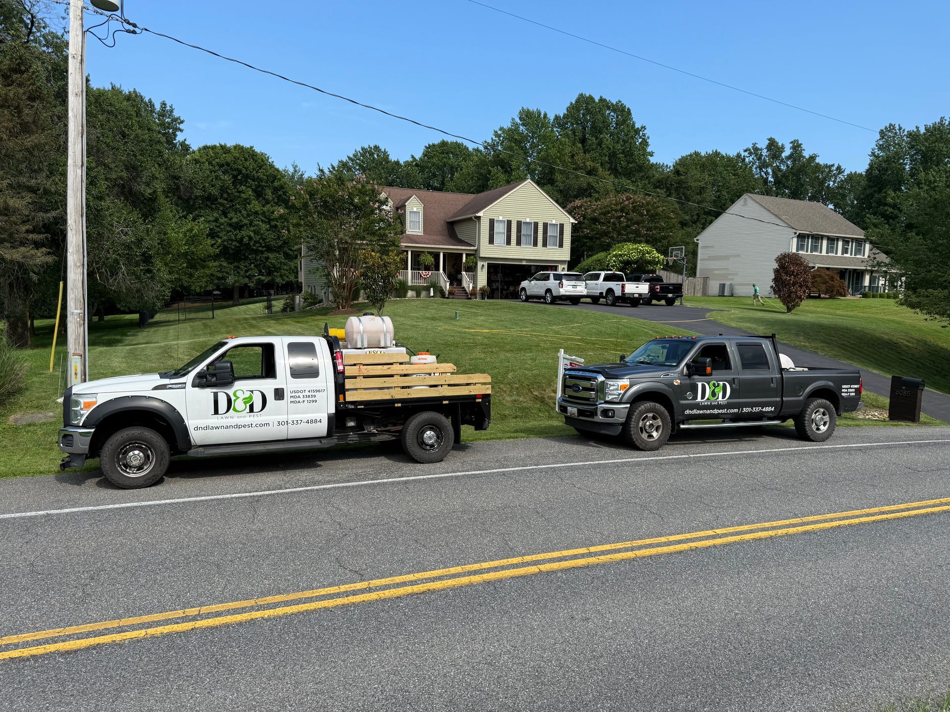 Two work trucks parked on a road in front of houses; one white, one gray, both with company logos.