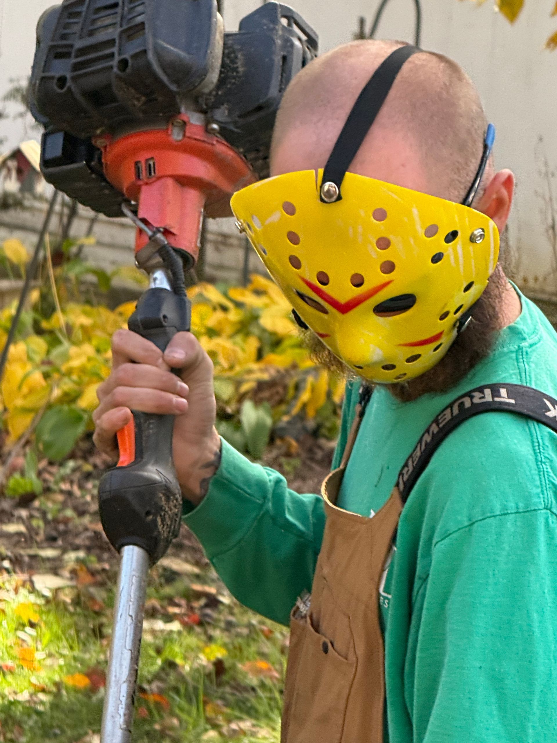 Man wearing a yellow hockey mask and using a weed whacker.
