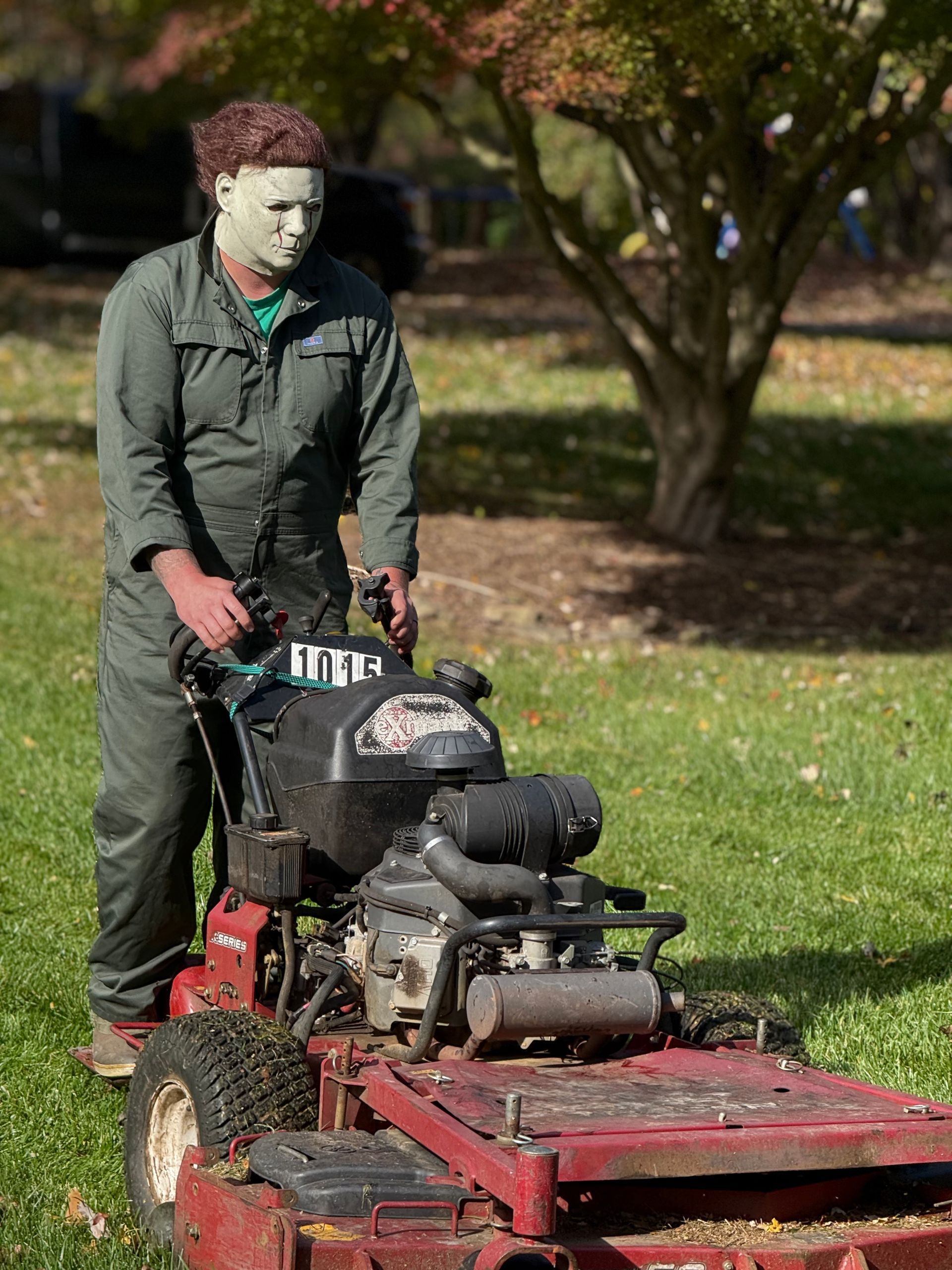 Person in a Michael Myers mask mowing grass with a red lawnmower on a sunny day.