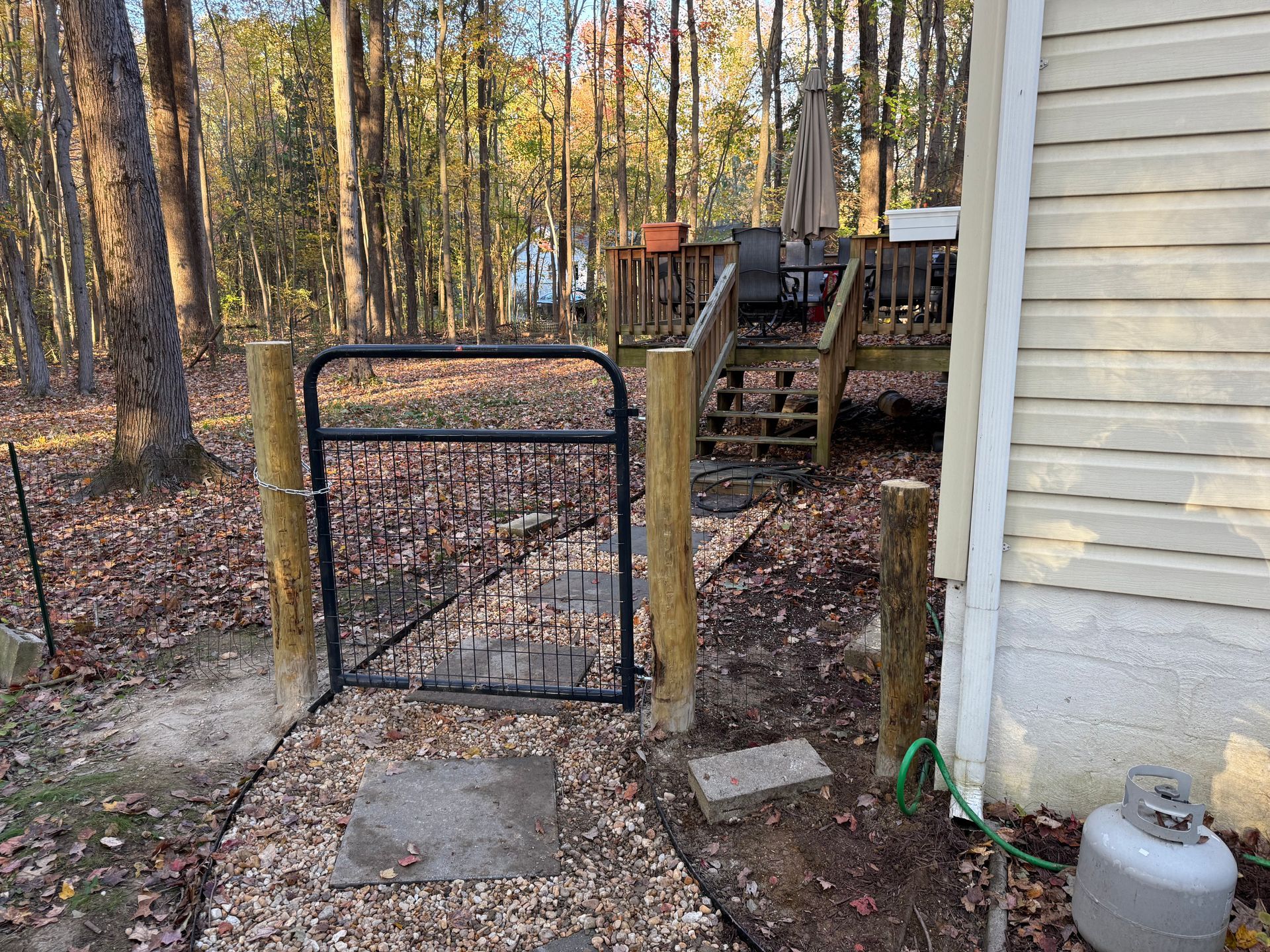 Black gate, wooden posts, path through wooded backyard, leading to deck.