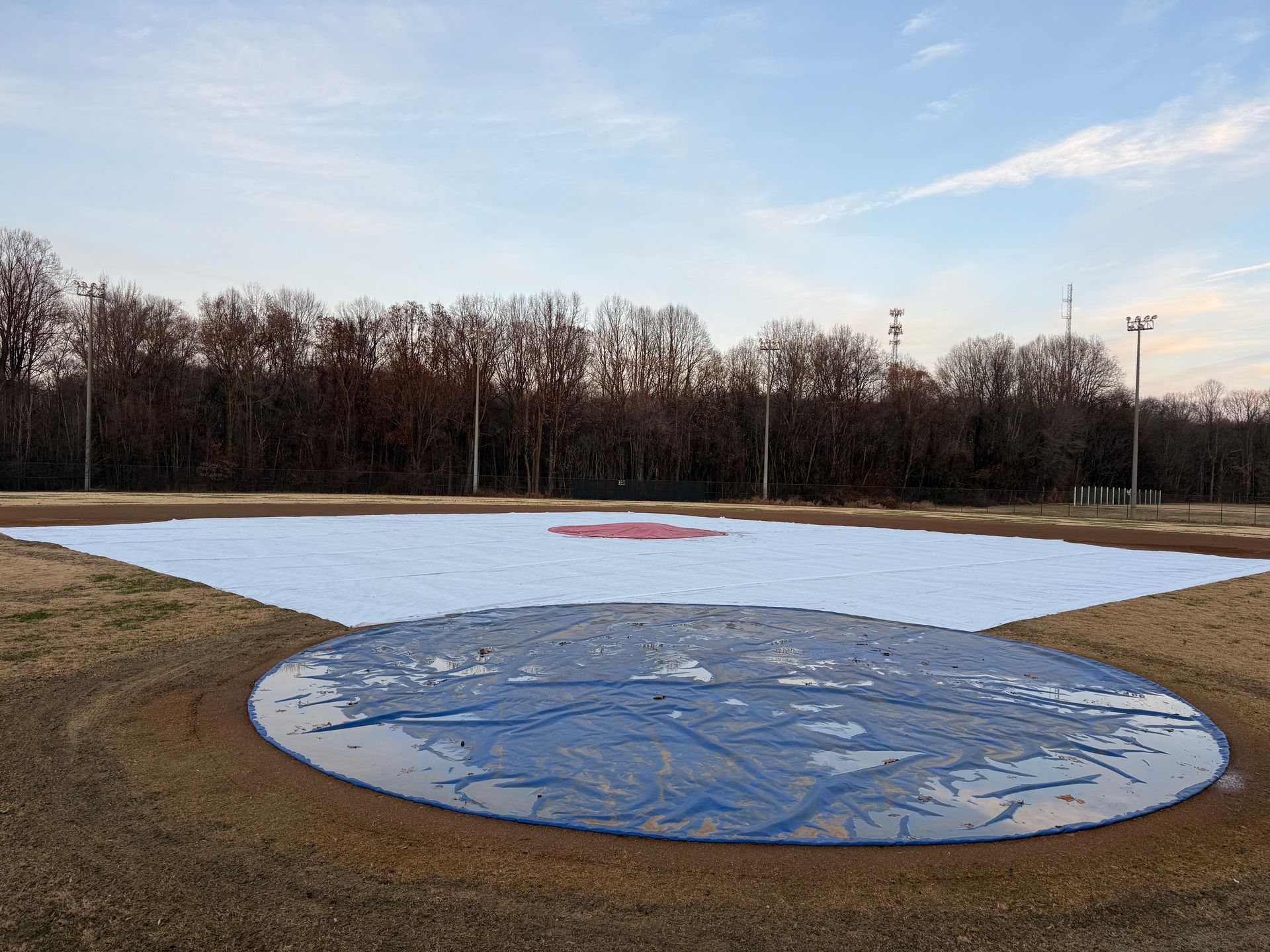 Baseball field with white and blue covers; trees in background. Cloudy sky.