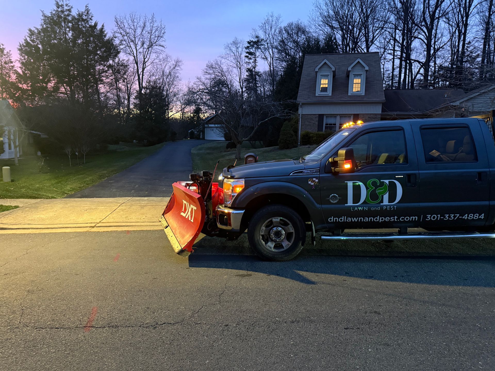Snowplow truck with red blade on a residential street at dusk.