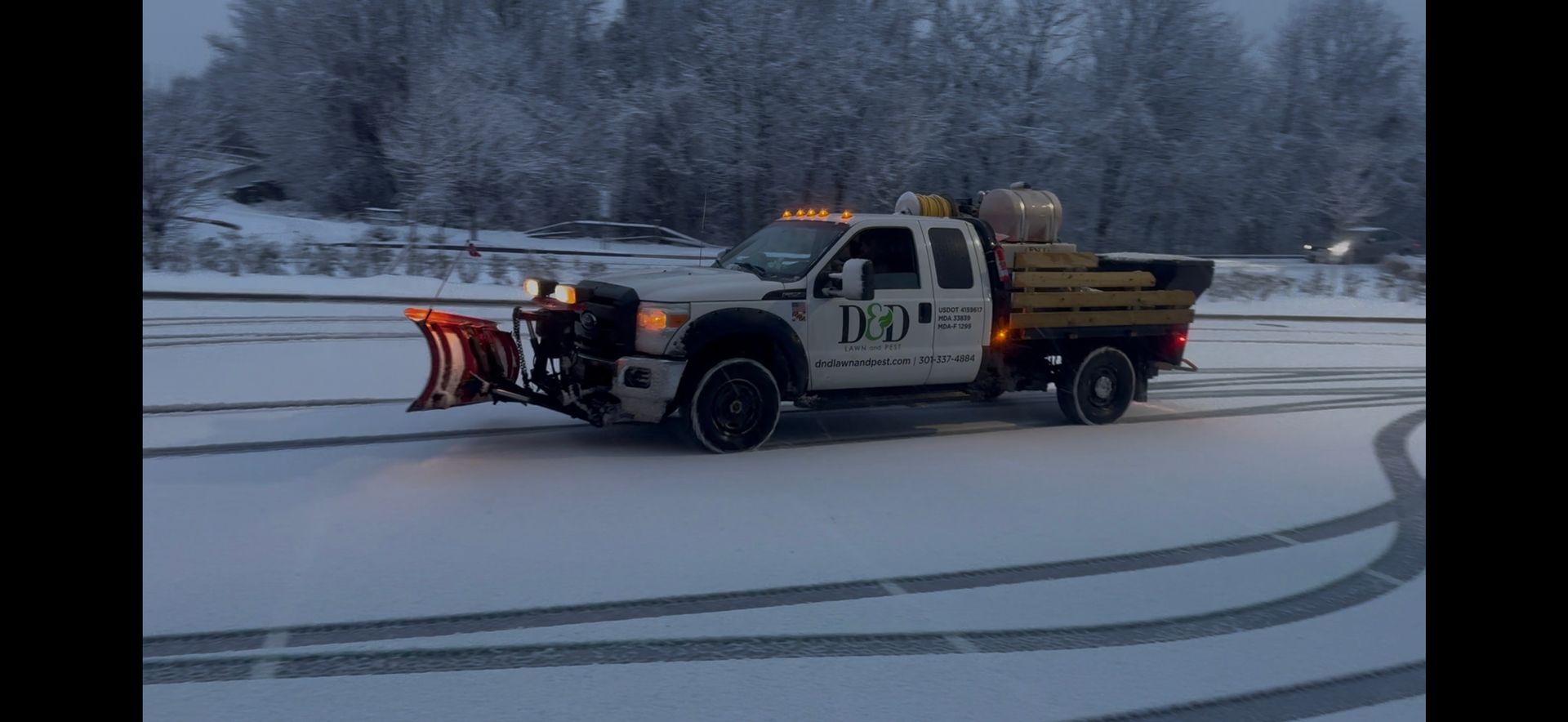 A snow plow truck clearing snow-covered road. Gray sky, trees in background, truck's lights are on.